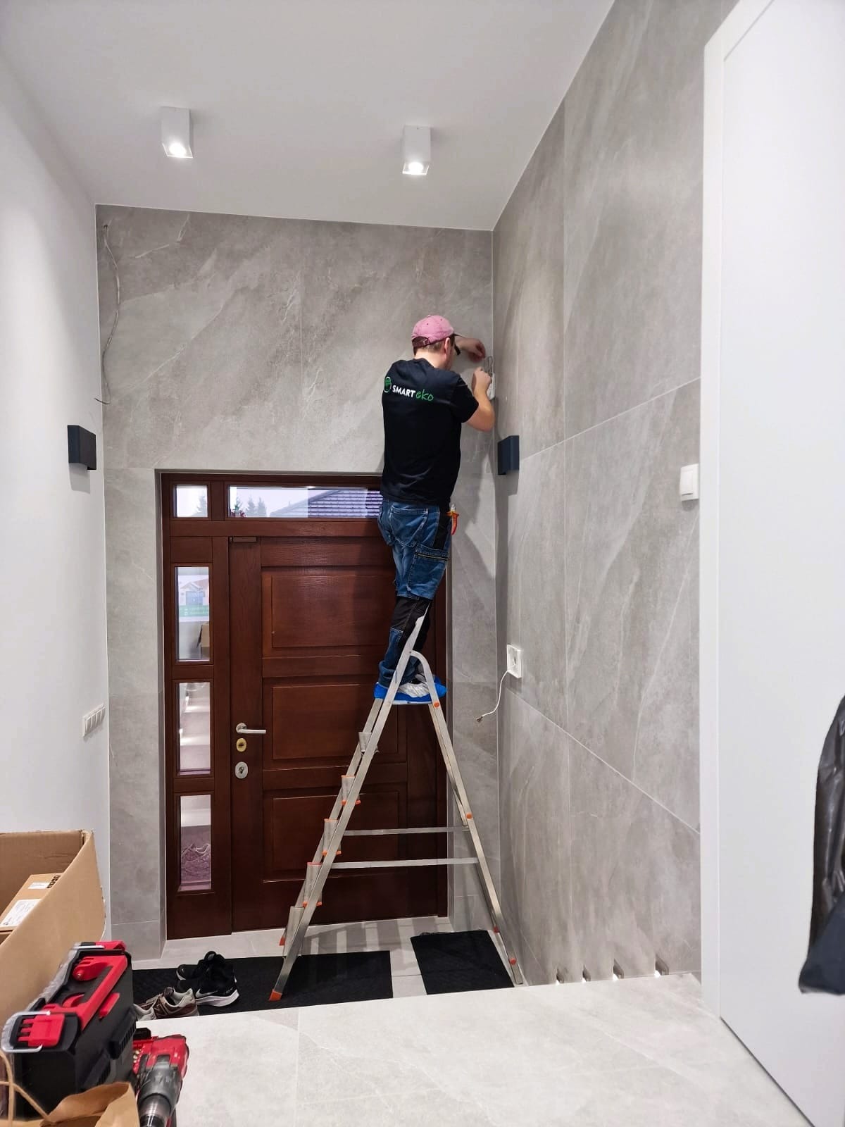 Elektryk na drabinie montuje czujnik alarmowy na marmurowej ścianie w korytarzu. Widoczna skrzynka narzędziowa i wejście do domu.
