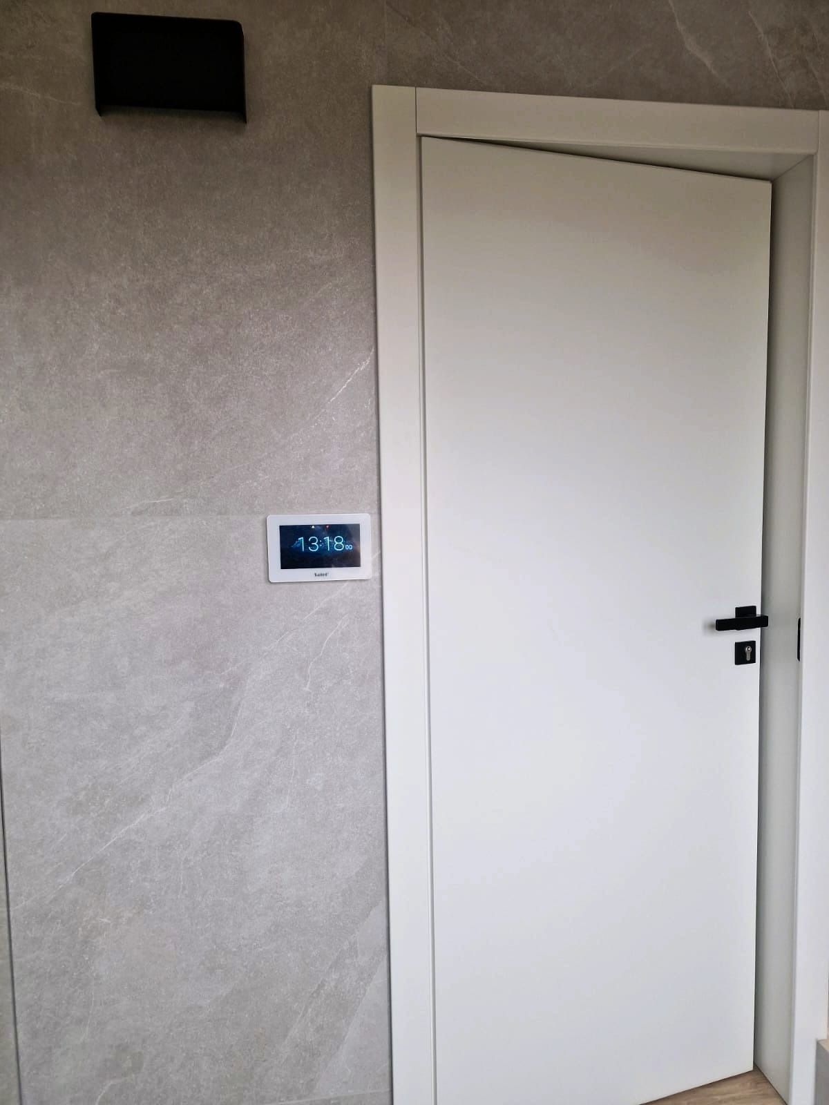 Panel sterowania inteligentnego domu na szarej ścianie obok białych drzwi z czarną klamką. Widoczny czujnik i oświetlenie.