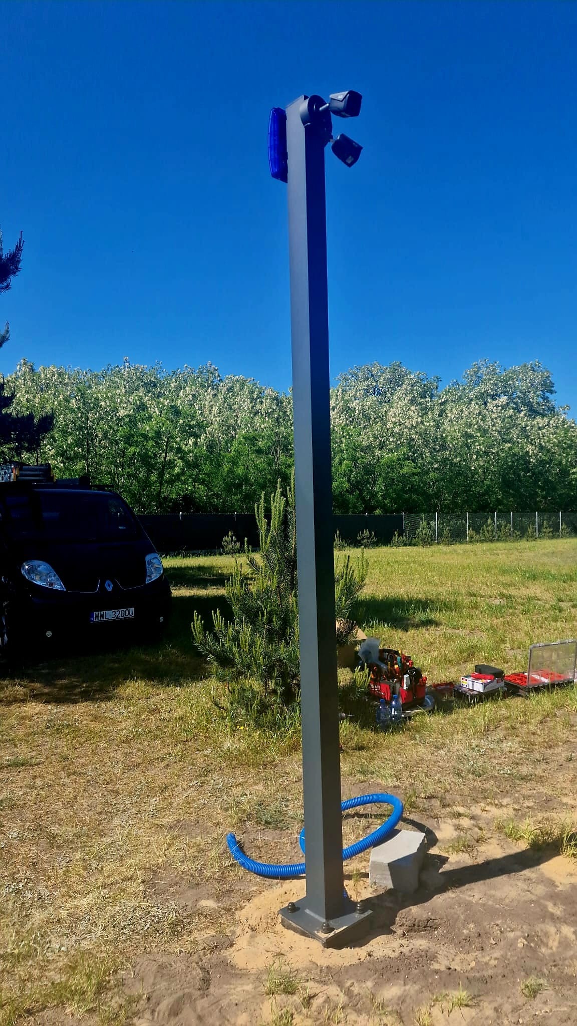 Słup monitoringu z kamerami i niebieskim światłem ostrzegawczym na tle błękitnego nieba i zieleni. Widoczne narzędzia i samochód w tle.