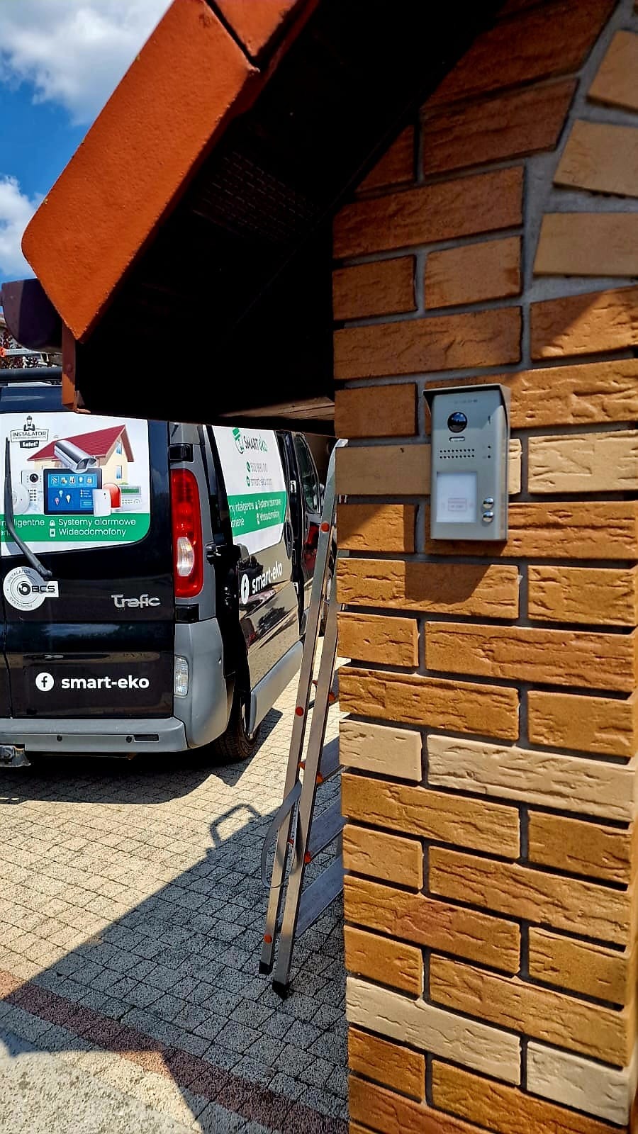 Szary wideodomofon zamontowany na ceglanej ścianie domu, obok drabina i samochód firmy Smart-Eko z reklamą systemów alarmowych i wideodomofonów.