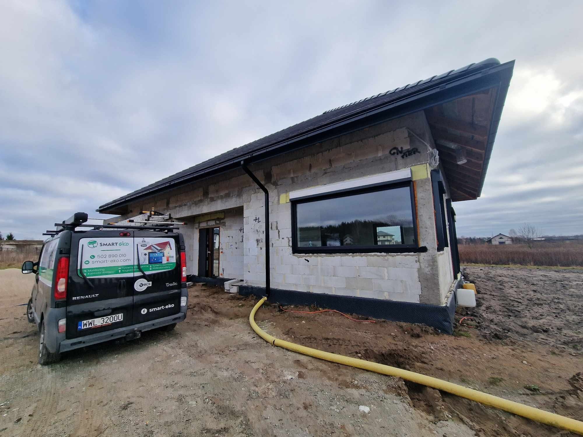 Surowy budynek z zamontowanymi oknami, widoczny samochód firmy Smart-Eko z drabiną na dachu i żółtym przewodem na ziemi. Budowa w toku, teren wokół rozkopany.