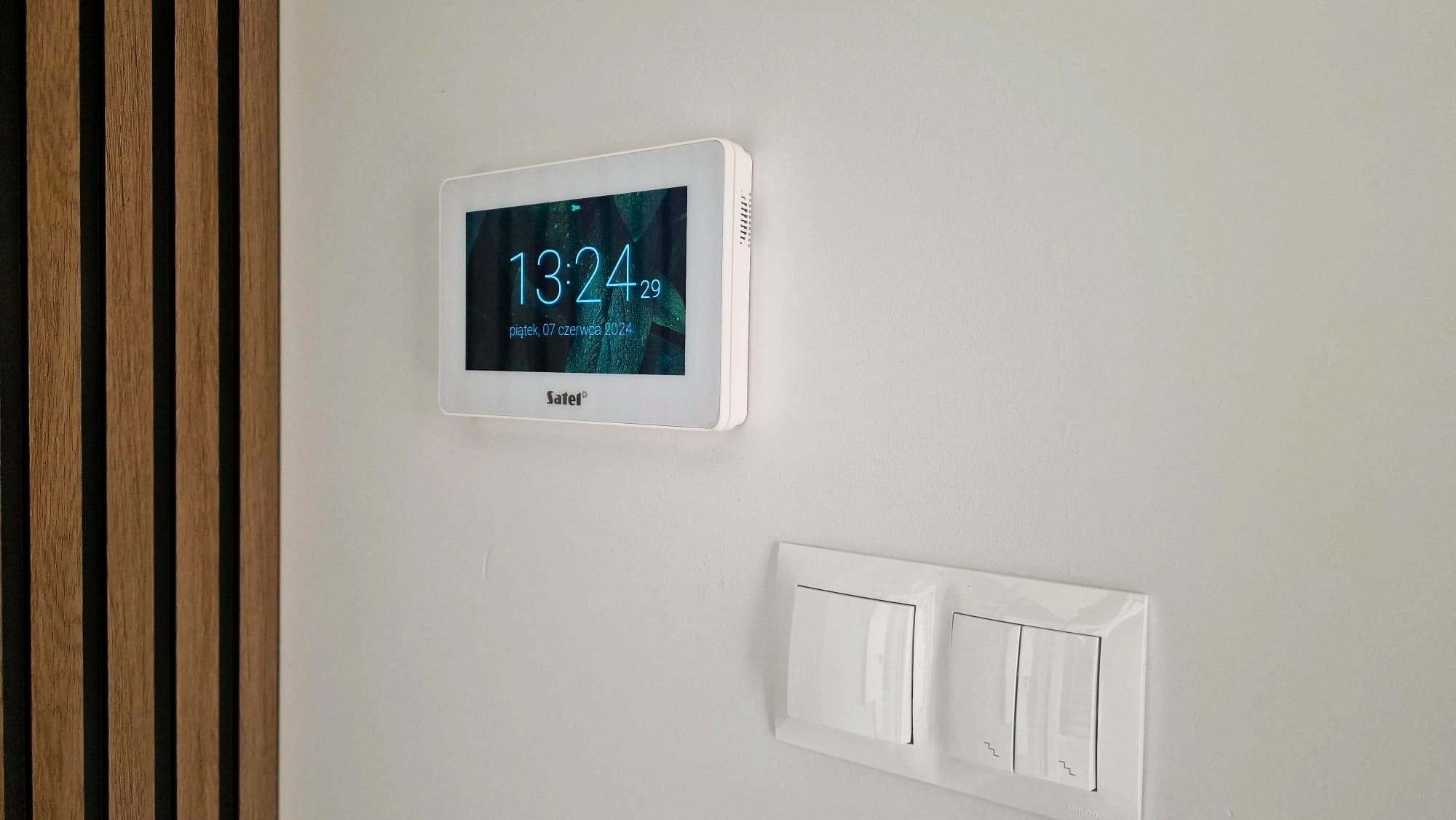 Biały panel dotykowy Safel z wyświetlaną godziną i datą, zamontowany na ścianie obok włącznika światła i dekoracyjnych paneli ściennych.
