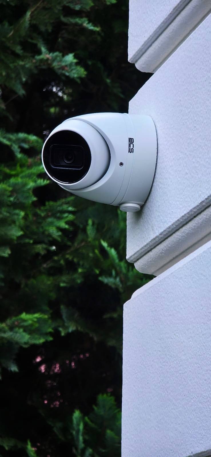 Kamera monitoringu BCS, kopułkowa, zamontowana na elewacji budynku z dekoracyjnym gzymsem. W tle zieleń drzew. Ujęcie z dołu.