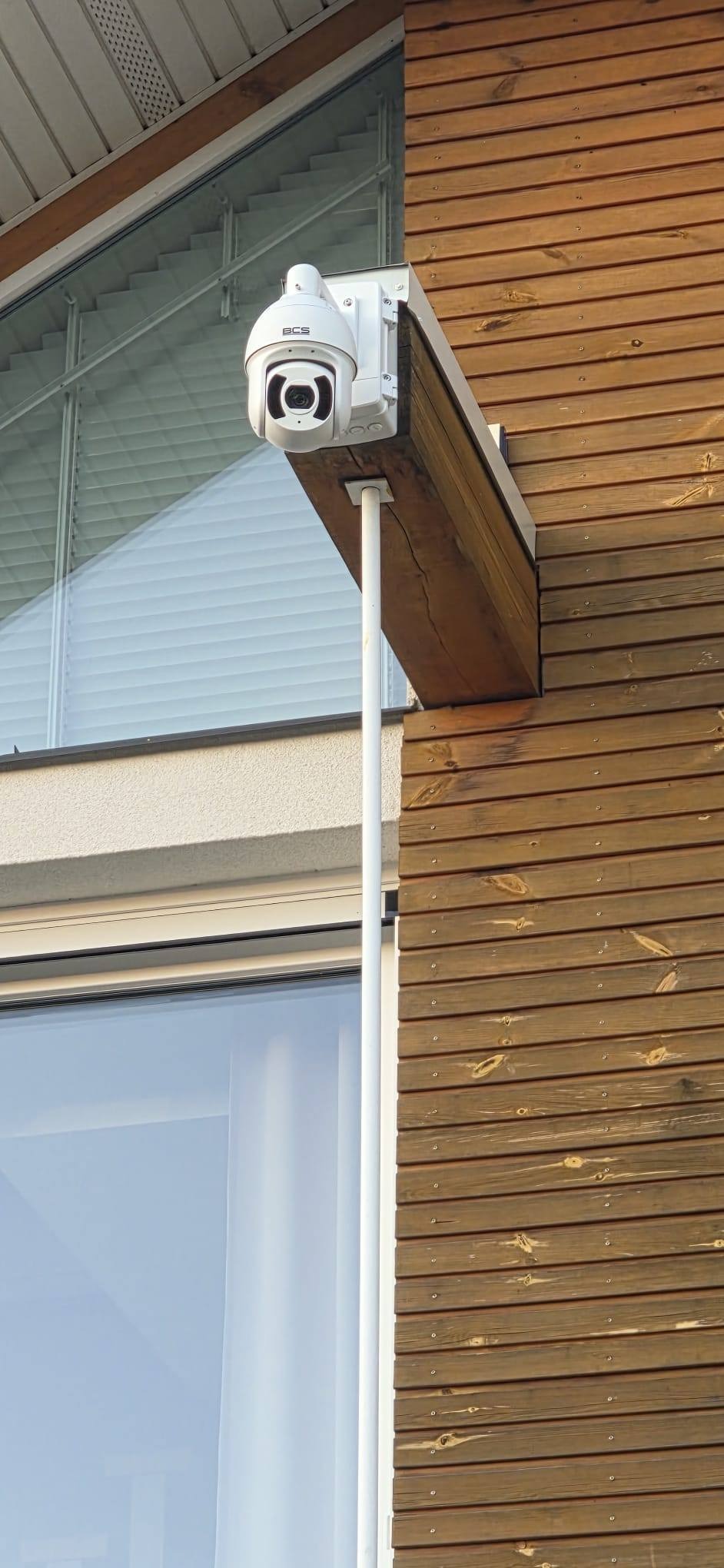 Kamera monitoringu BCS zamontowana na drewnianej elewacji budynku, widoczna z boku, z oknem w tle. Biała obudowa kamery kontrastuje z brązowym drewnem.