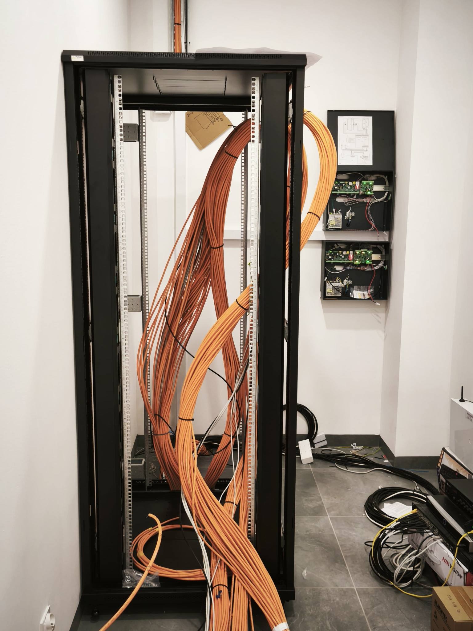 Szafa rack z pomarańczowymi kablami w trakcie instalacji. Na ścianie skrzynka z elektroniką. Widoczne przewody i elementy montażowe. Szara podłoga.