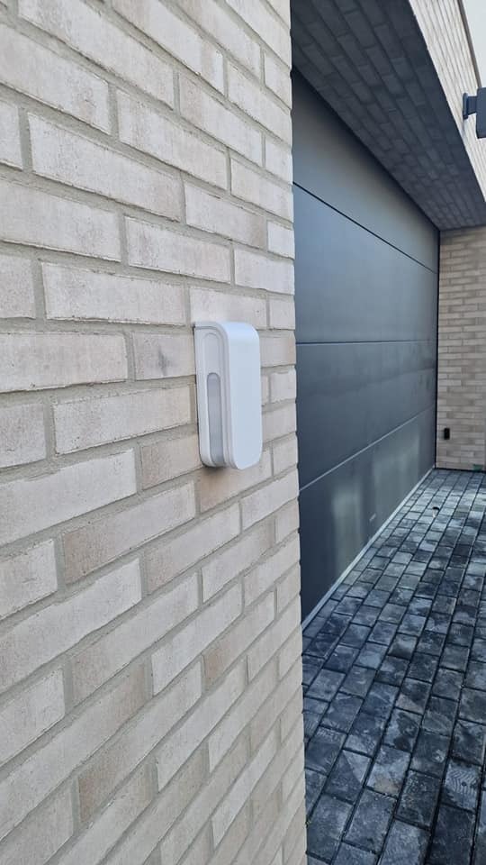 Biały czujnik ruchu zamontowany na ceglanej ścianie obok nowoczesnej, szarej bramy garażowej. Widok z boku, oświetlenie dzienne.