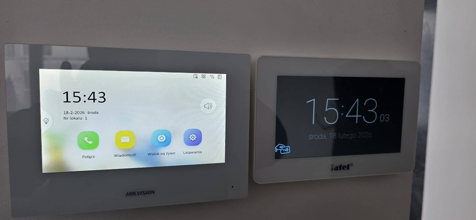 Dwa panele wideodomofonowe różnych producentów (Hikvision i Satel) zamontowane obok siebie na ścianie, wyświetlające aktualną godzinę i datę.