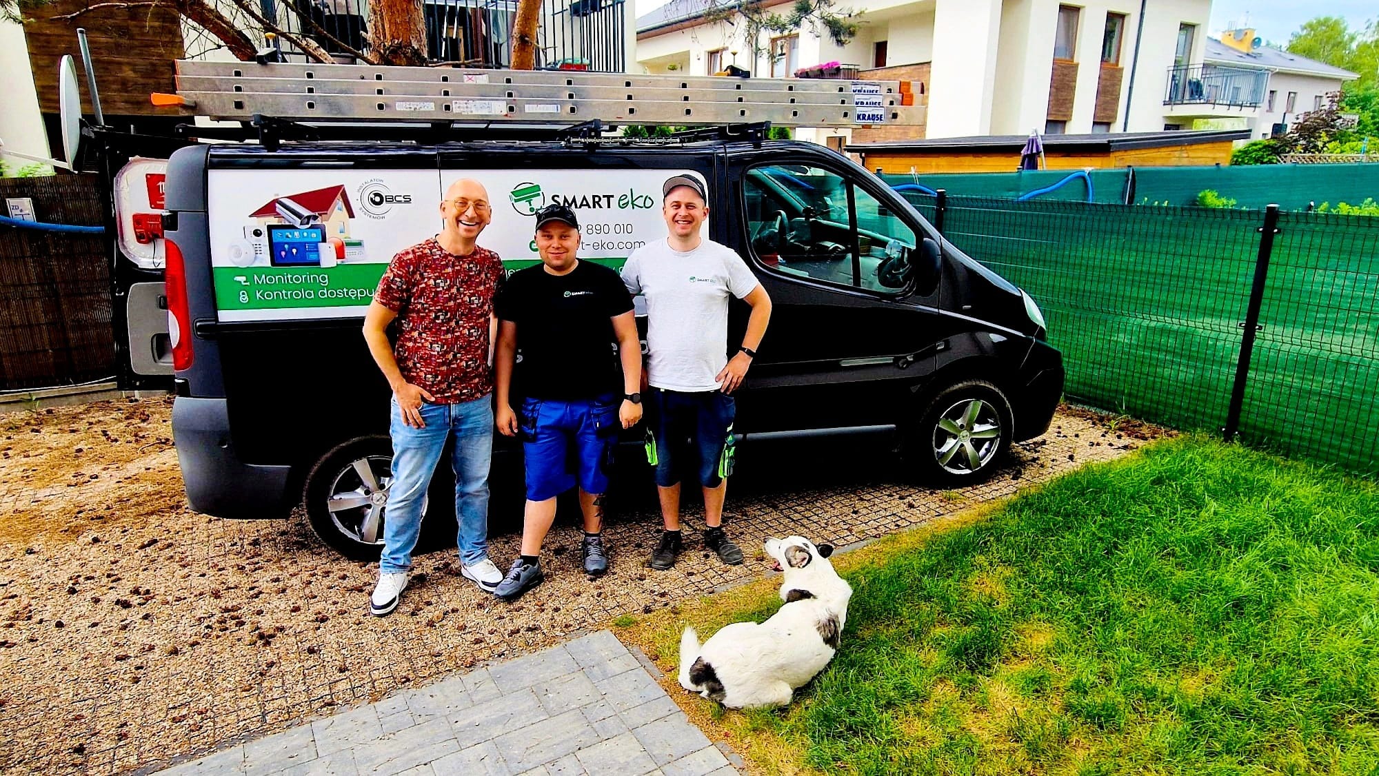 Ekipa SMART eko prezentuje firmowy van z drabiną, monitoring i kontrola dostępu. Uśmiechnięci instalatorzy i pies na tle domu i zielonego ogrodzenia.