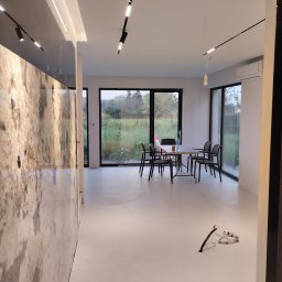 Piotr Kowalski - Nowoczesne wnętrze z marmurową ścianą, minimalistycznym oświetleniem i widokiem na zieleń za oknem. Biała podłoga i czarne krzesła dodają kontrastu.