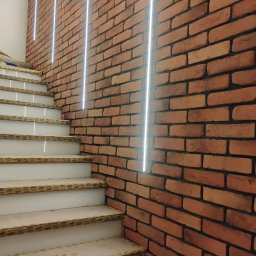 Piotr Kowalski - Klatka schodowa z ceglaną ścianą i oświetleniem LED. Widoczne zabezpieczone schody podczas prac wykończeniowych. Cegła w odcieniach czerwieni i brązu.