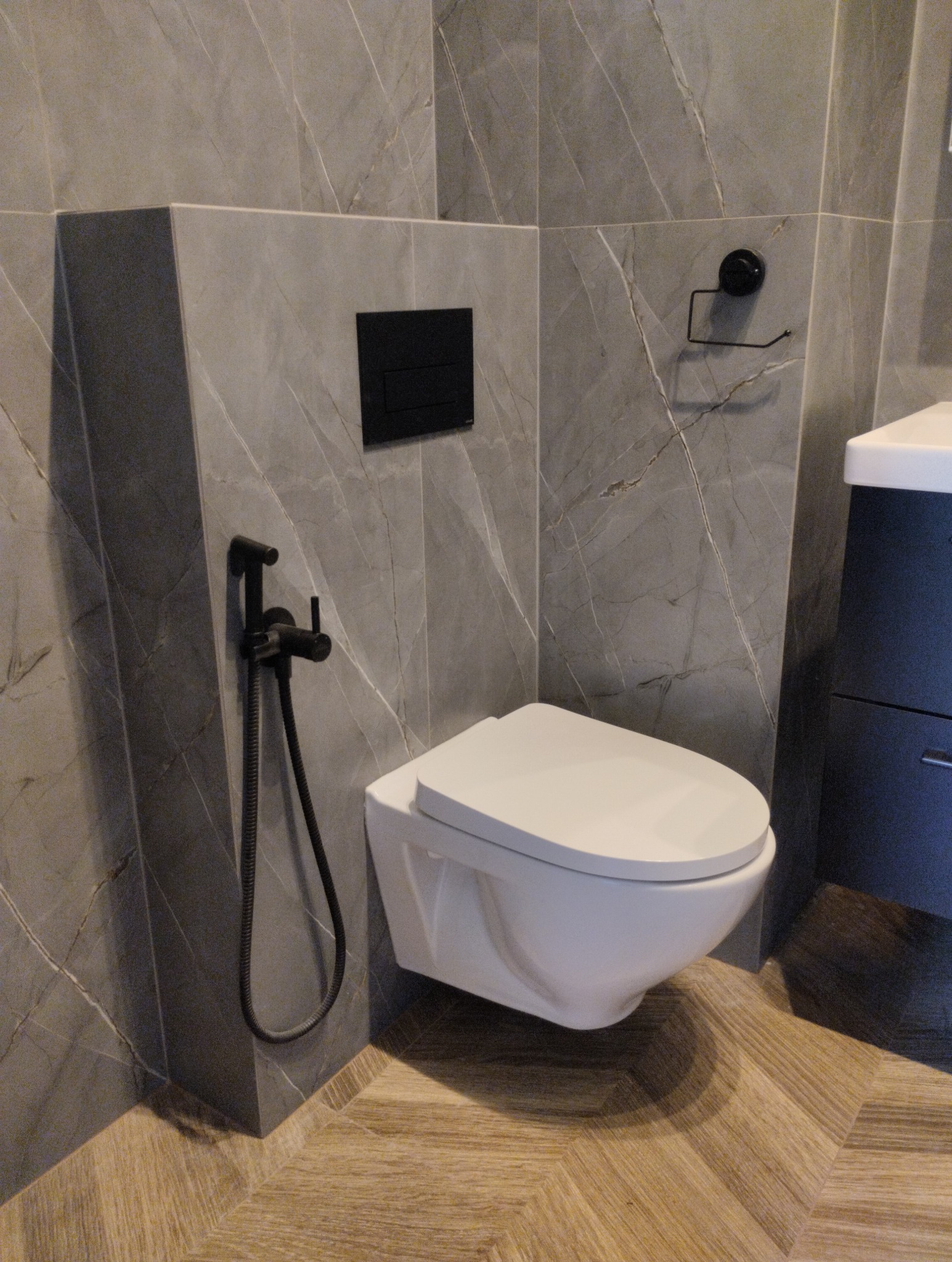 Nowoczesna toaleta podwieszana z czarną armaturą i szarą marmurową okładziną ścienną. Uwagę zwraca parkiet ułożony w jodełkę francuską.