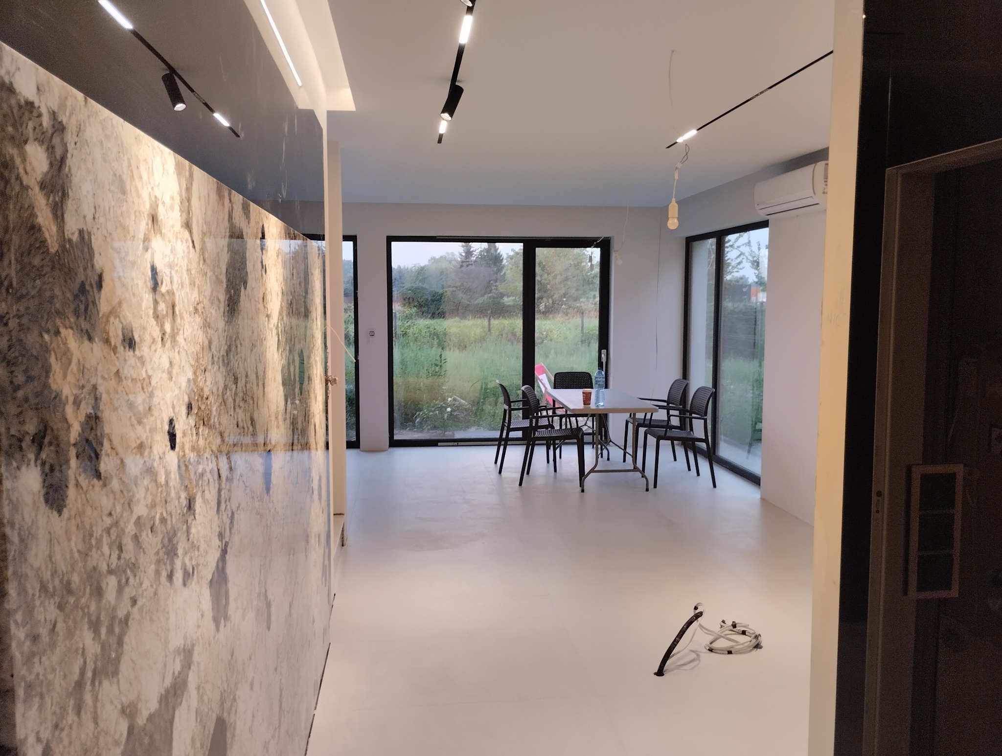 Nowoczesne wnętrze z marmurową ścianą, minimalistycznym oświetleniem i widokiem na zieleń za oknem. Biała podłoga i czarne krzesła dodają kontrastu.