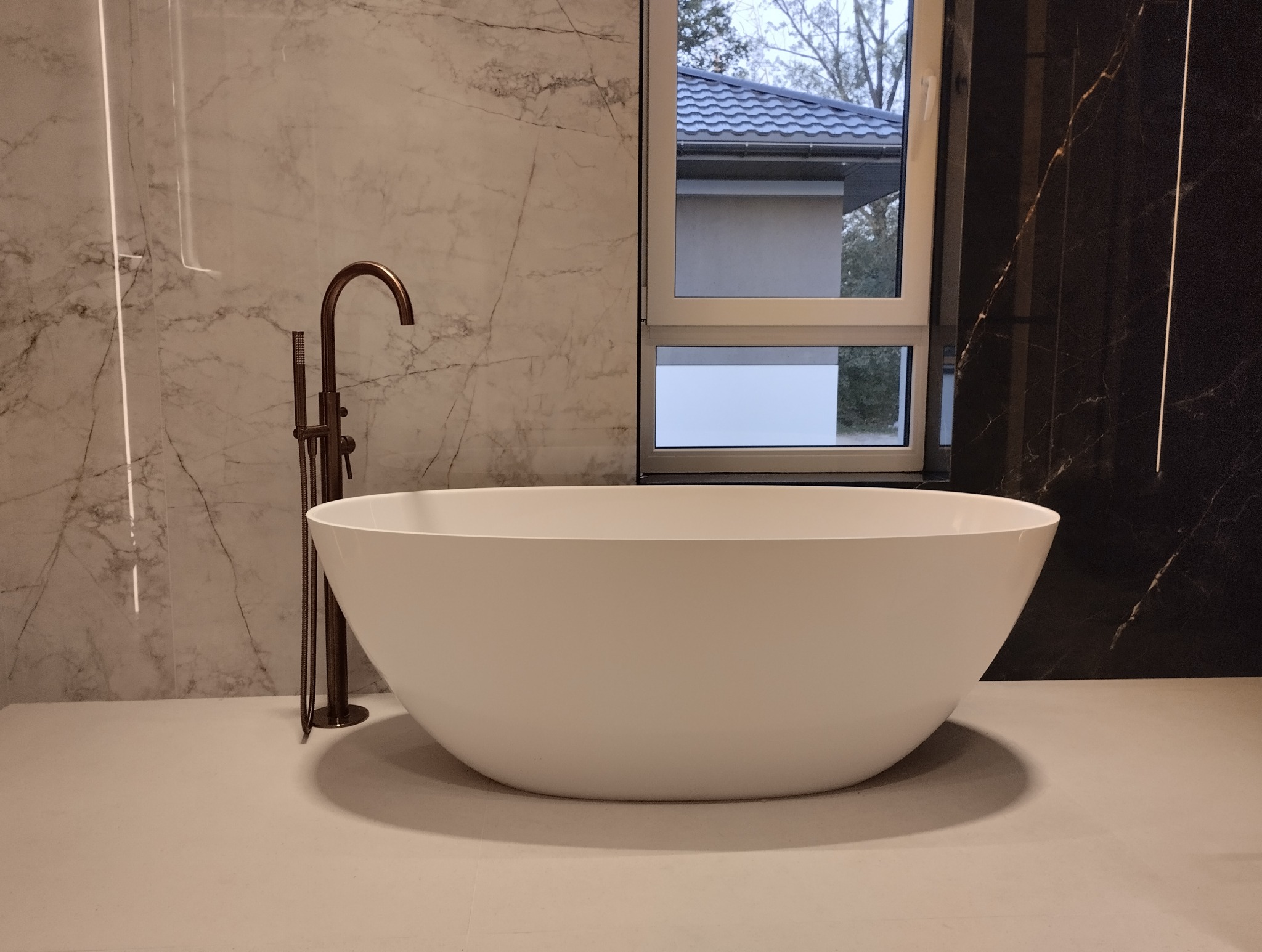 Biała, wolnostojąca wanna z brązową armaturą na tle marmurowych ścian i okna z widokiem na zewnątrz. Nowoczesna aranżacja łazienki z akcentem na naturalne materiały.