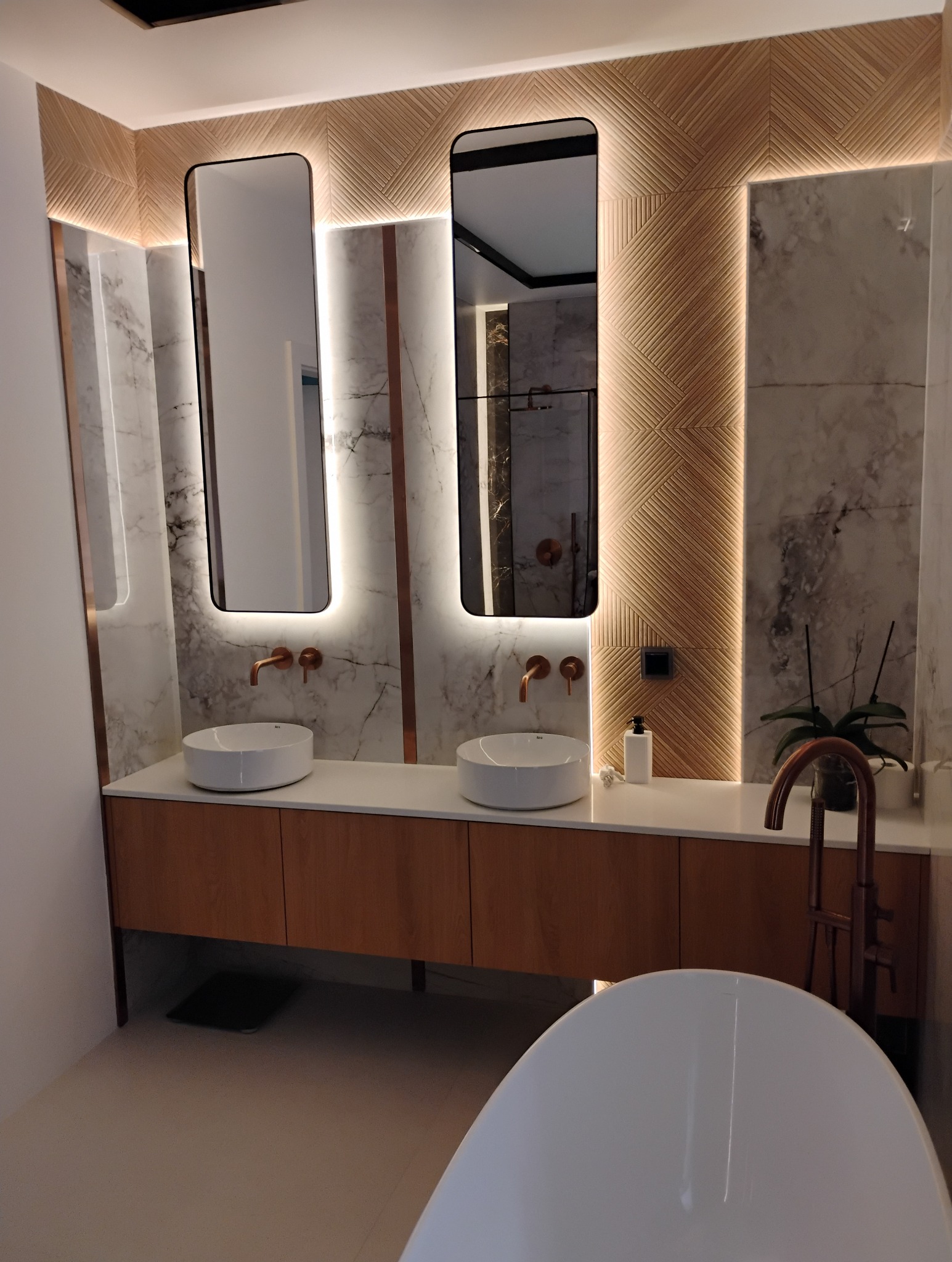 Nowoczesna łazienka z dwoma umywalkami, podświetlanymi lustrami i drewnianą zabudową. Marmurowe ściany i dekoracyjne panele ścienne tworzą elegancki wystrój.