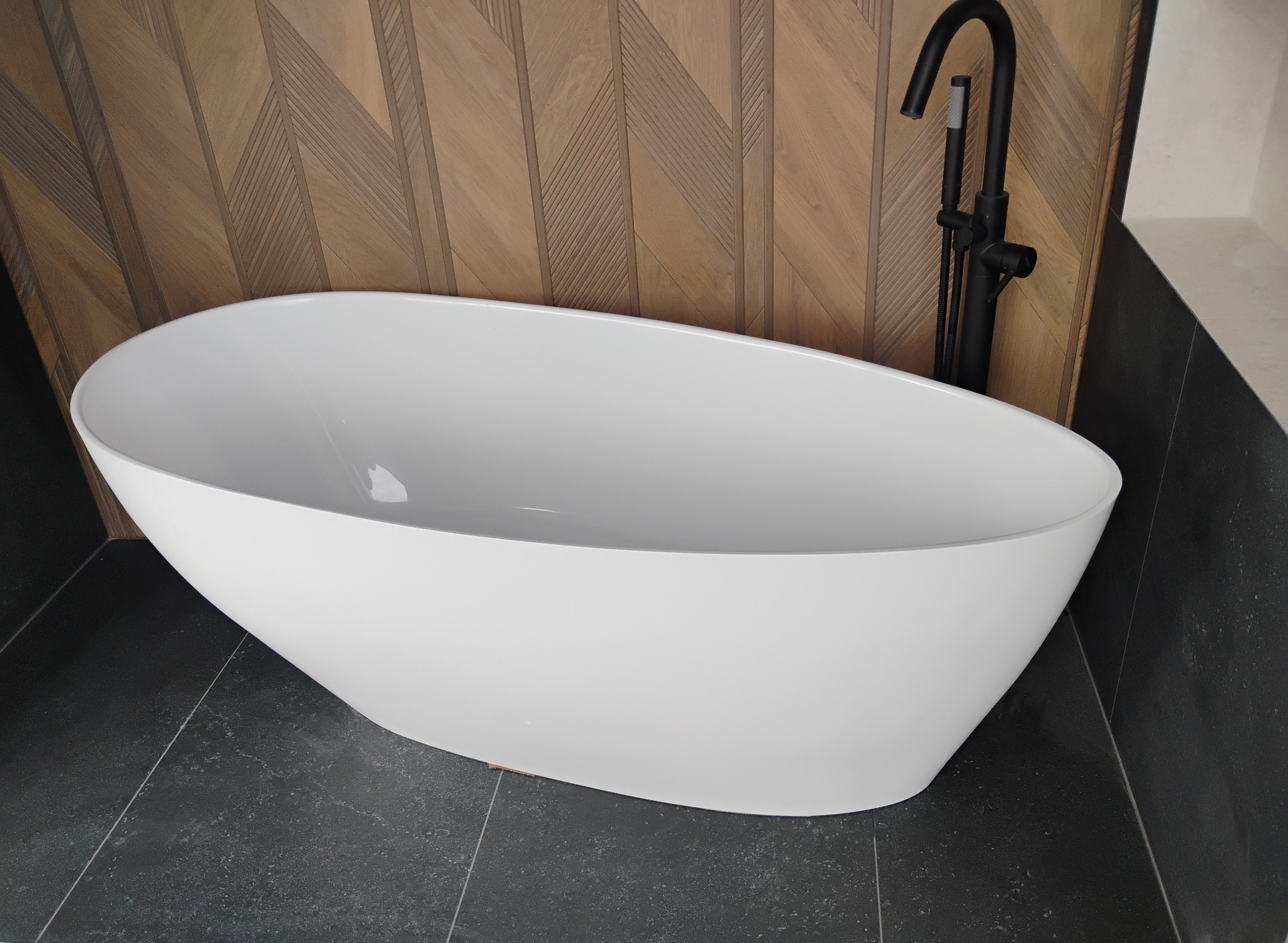 Nowoczesna, wolnostojąca wanna w łazience po wykończeniu w Zielonce. Biała armatura na tle drewnianej ściany i ciemnej podłogi z płytek. Minimalistyczny design.