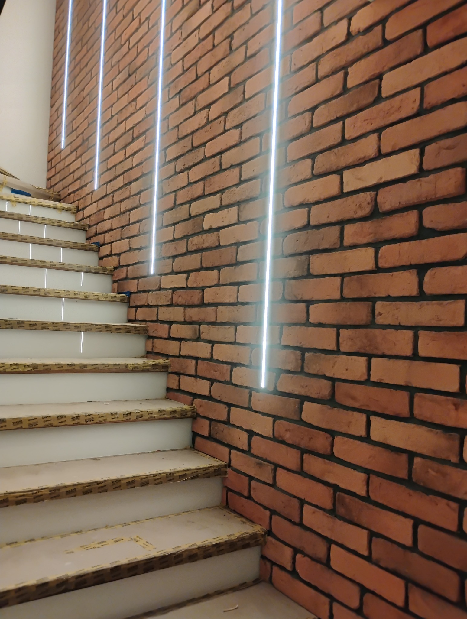Klatka schodowa z ceglaną ścianą i oświetleniem LED. Widoczne zabezpieczone schody podczas prac wykończeniowych. Cegła w odcieniach czerwieni i brązu.