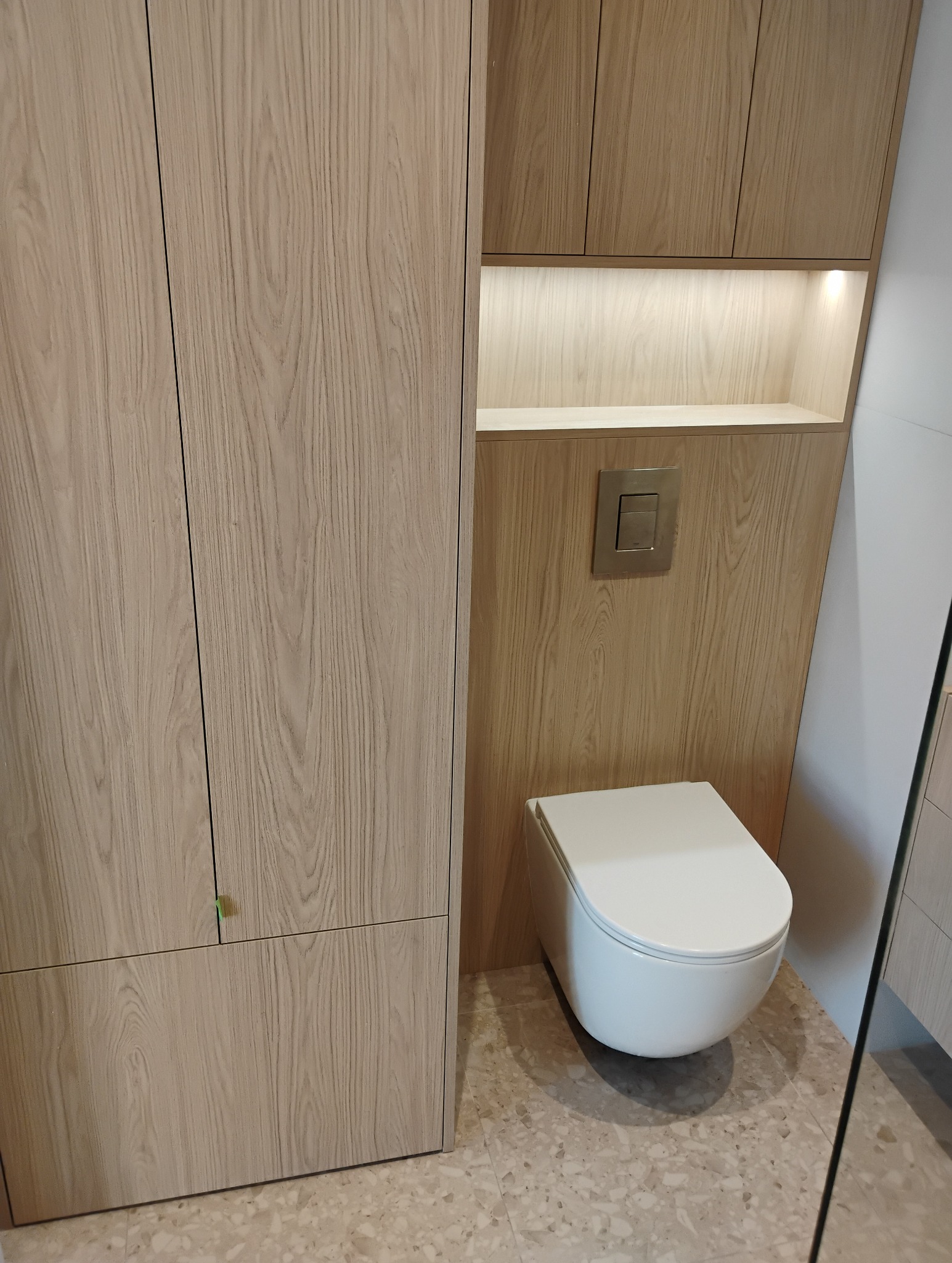 Nowoczesna toaleta podwieszana w zabudowie z drewnopodobnej płyty, z szafką i podświetlaną wnęką. Minimalistyczny design, beżowa kolorystyka, płytki lastryko na podłodze.