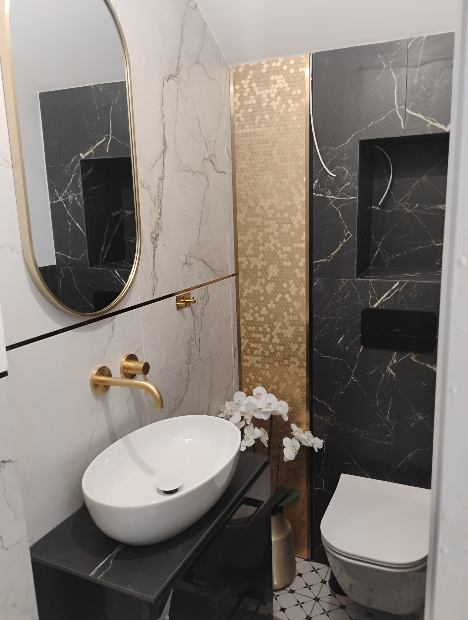 Elegancka łazienka po remoncie: biała umywalka nablatowa, złota armatura, lustro w złotej ramie, płytki imitujące marmur i mozaika. Nowoczesny design.