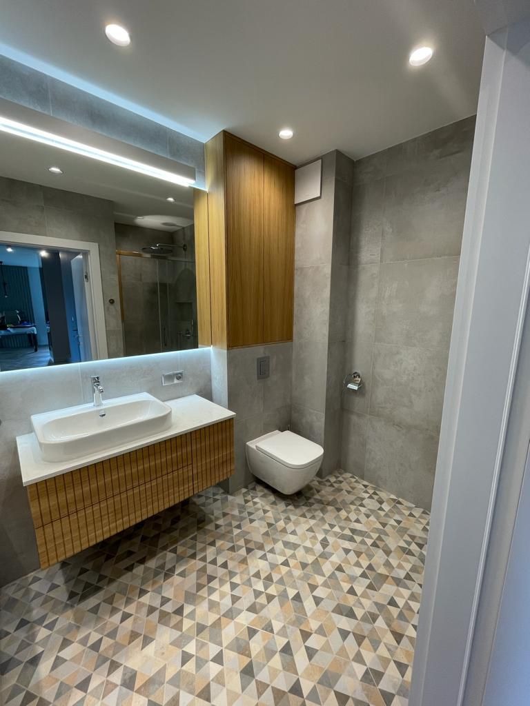 Nowoczesna łazienka z drewnianą szafką pod umywalką, geometryczną podłogą i szarą glazurą. Lustro z podświetleniem LED i wisząca toaleta.