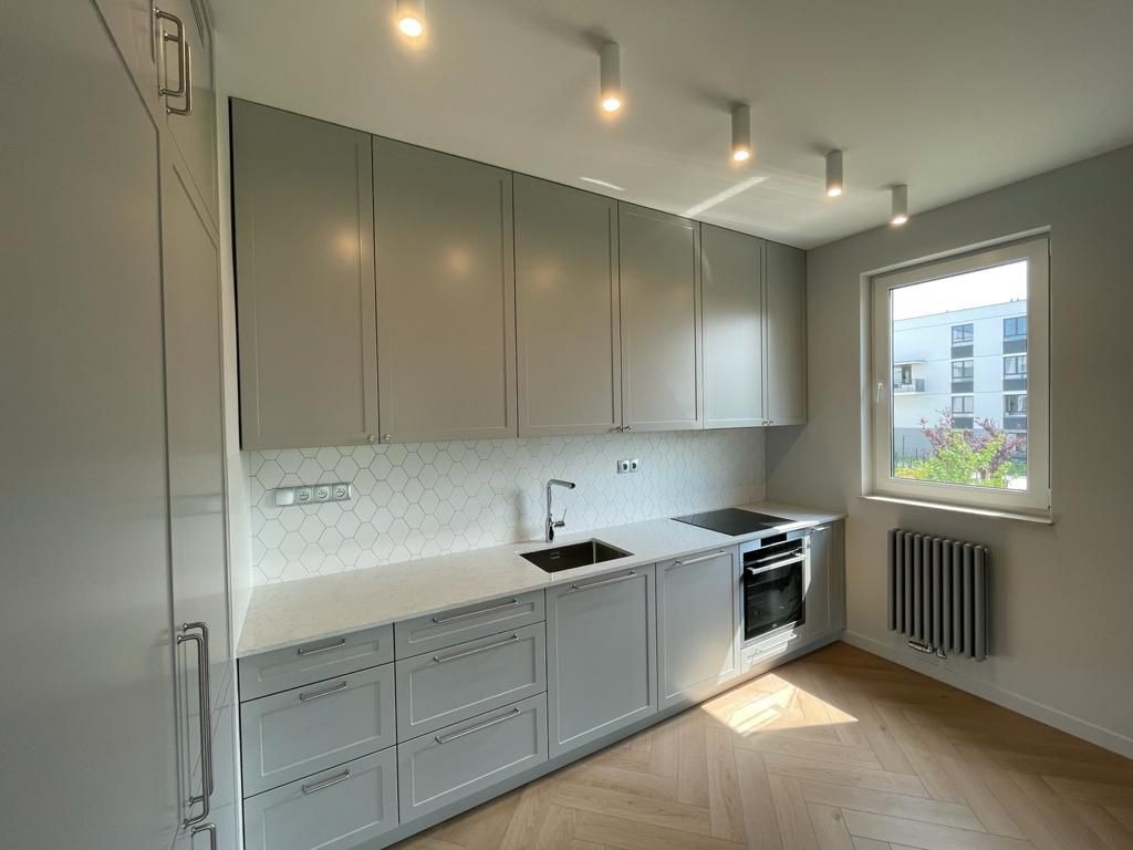 Jasnoszara kuchnia z białym blatem i heksagonalnymi płytkami. Parkiet ułożony w jodełkę. Nowoczesne oświetlenie punktowe i grzejnik pod oknem.