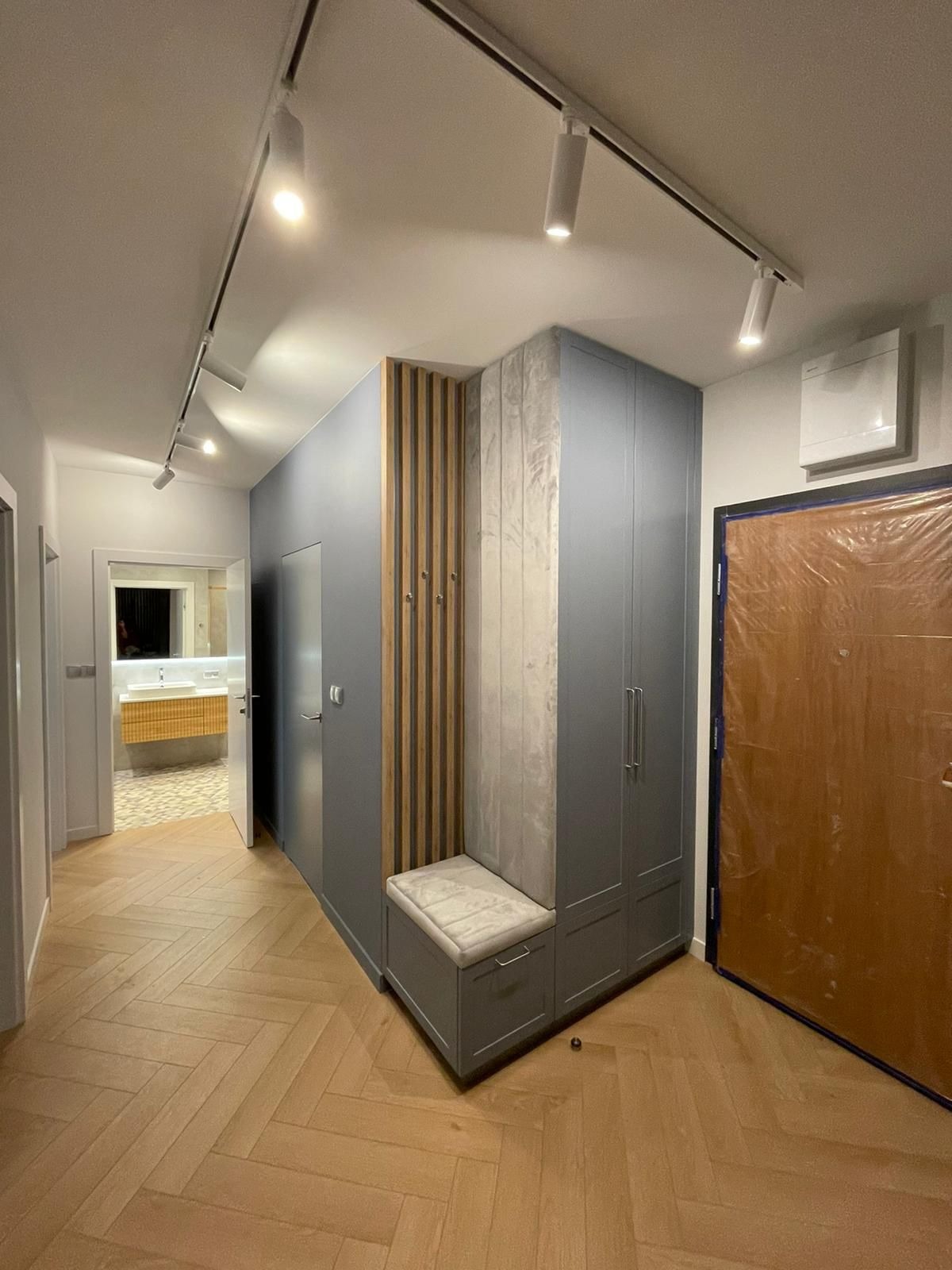 Nowoczesny przedpokój z szafą w zabudowie, drewnianymi lamelami i siedziskiem. Na podłodze parkiet ułożony w jodełkę. Widok na łazienkę w tle.