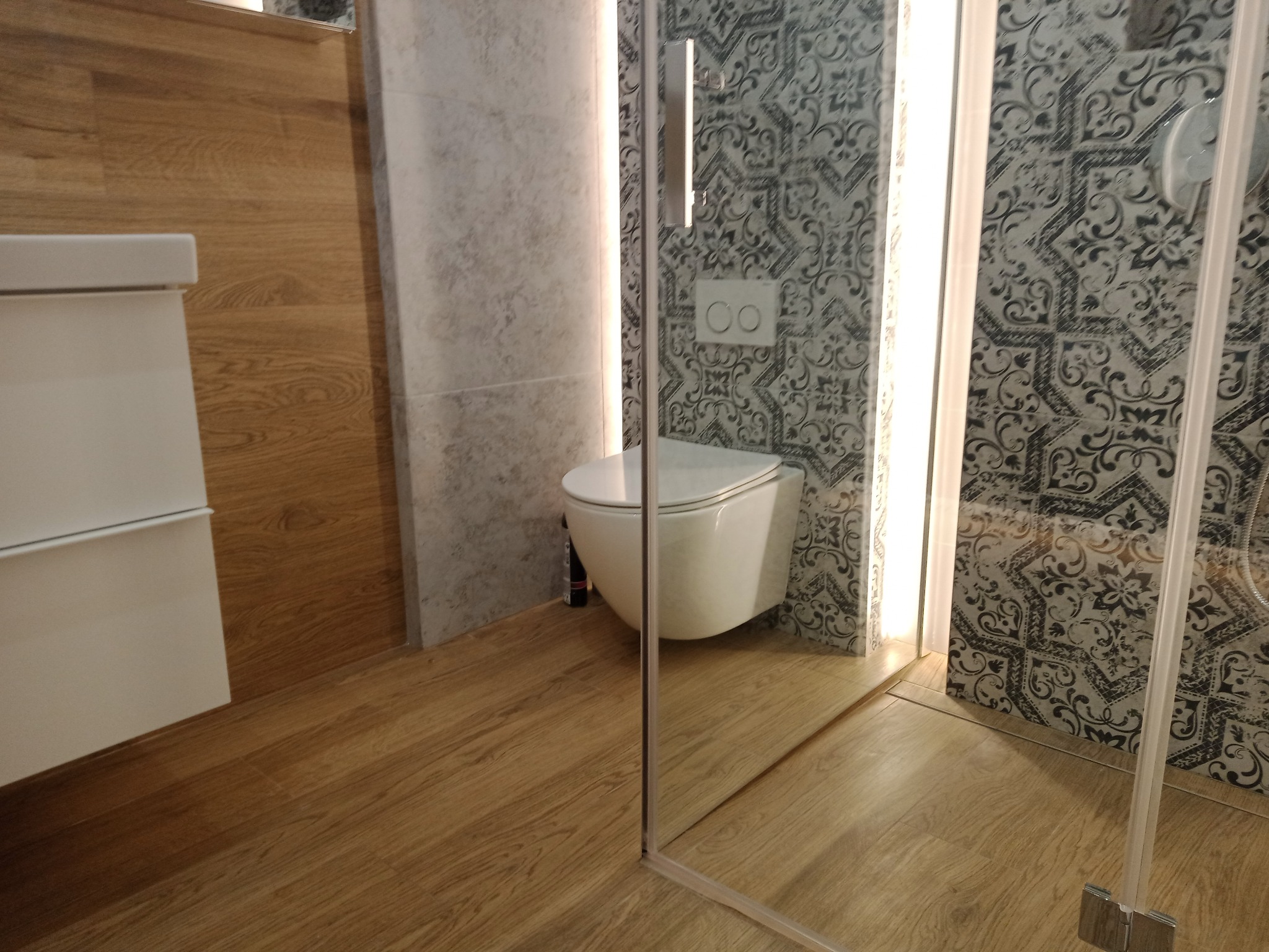 Nowoczesna łazienka po remoncie: wisząca toaleta, drewnopodobne płytki podłogowe i ścienne, kabina prysznicowa ze szklanymi drzwiami, wzorzyste płytki na ścianie.