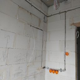 Usługi Elektryczne Przemysław Sekunda - Wewnętrzne okablowanie elektryczne w surowym budynku z białych bloków, widoczne kable w peszlach i pomarańczowe puszki instalacyjne. Na ścianach widoczne odręczne notatki.