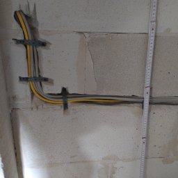 Usługi Elektryczne Przemysław Sekunda - Kable elektryczne w żółtej i szarej izolacji, przymocowane metalowymi uchwytami do surowej ściany z betonu komórkowego. Obok pionowo ustawiona miarka.