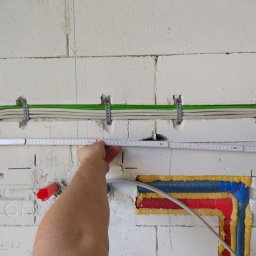 Usługi Elektryczne Przemysław Sekunda - Białe kable w zielonej osłonie, przymocowane do ściany z betonu komórkowego za pomocą metalowych uchwytów. Mierzone odległości taśmą mierniczą.
