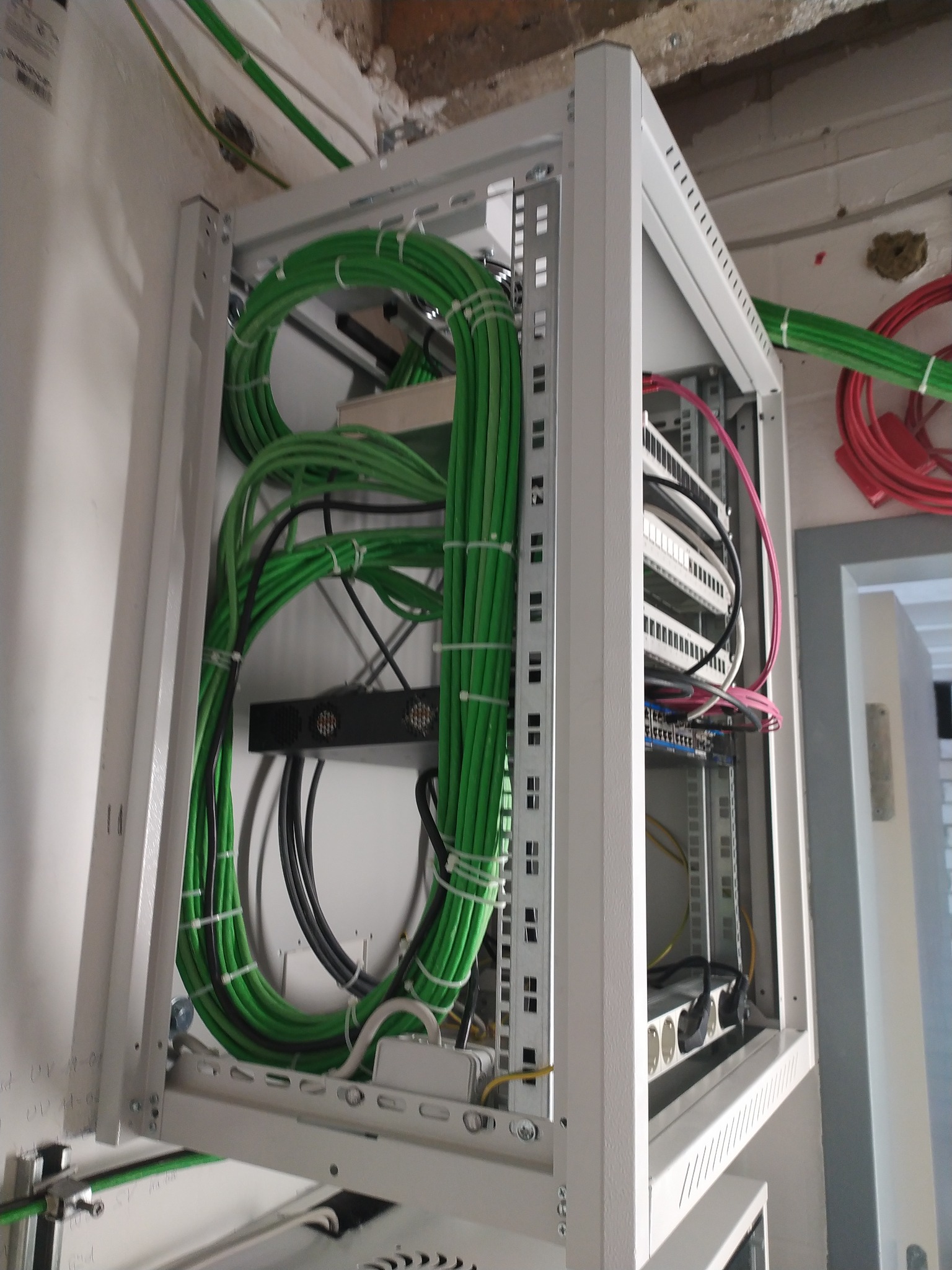 Szafa rack z uporządkowanymi kablami w kolorze zielonym i czarnym, zabezpieczonymi opaskami. Widoczne elementy montażowe i urządzenia wewnątrz szafy.