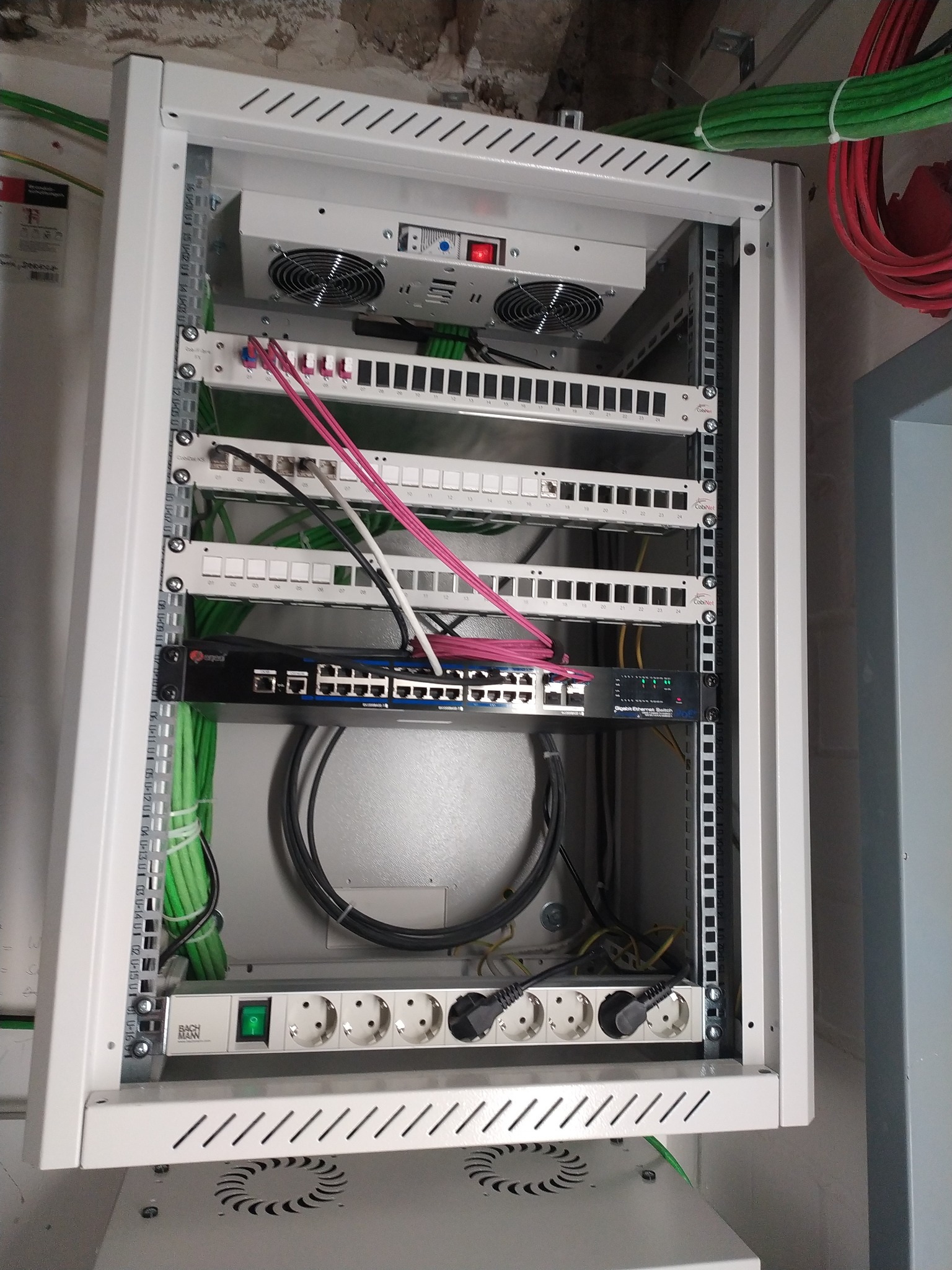 Szafa rack z zamontowanym switchem, patch panelem i listwą zasilającą. Widoczne kable w kolorach zielonym, czerwonym i czarnym. Górna część szafy z wentylatorami.