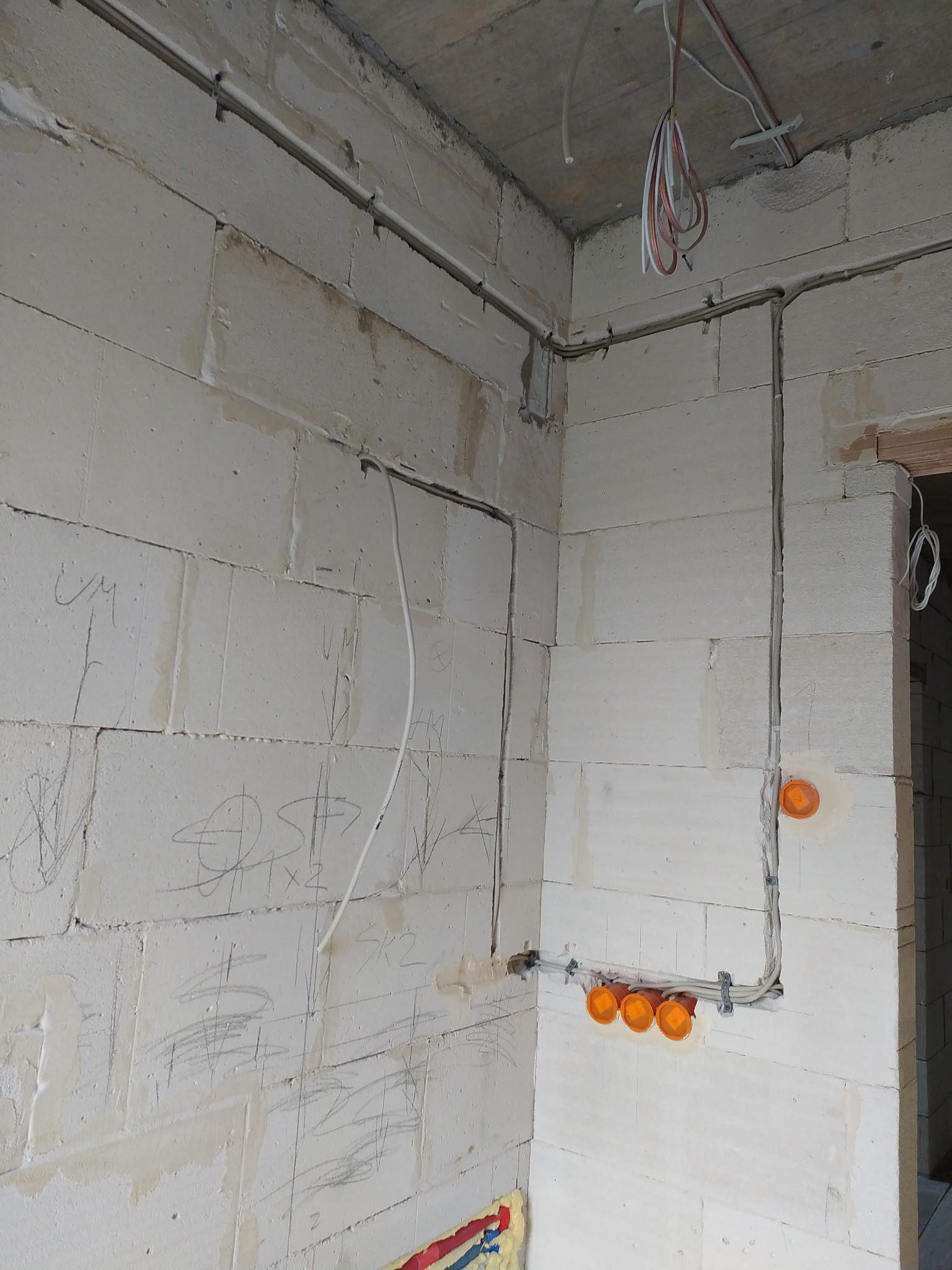 Wewnętrzne okablowanie elektryczne w surowym budynku z białych bloków, widoczne kable w peszlach i pomarańczowe puszki instalacyjne. Na ścianach widoczne odręczne notatki.