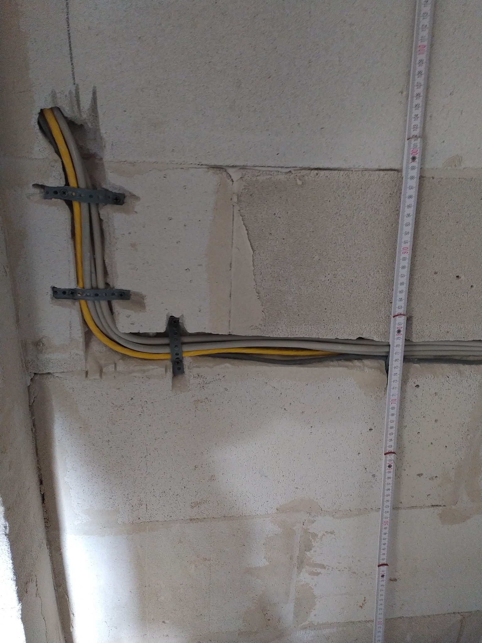Kable elektryczne w żółtej i szarej izolacji, przymocowane metalowymi uchwytami do surowej ściany z betonu komórkowego. Obok pionowo ustawiona miarka.