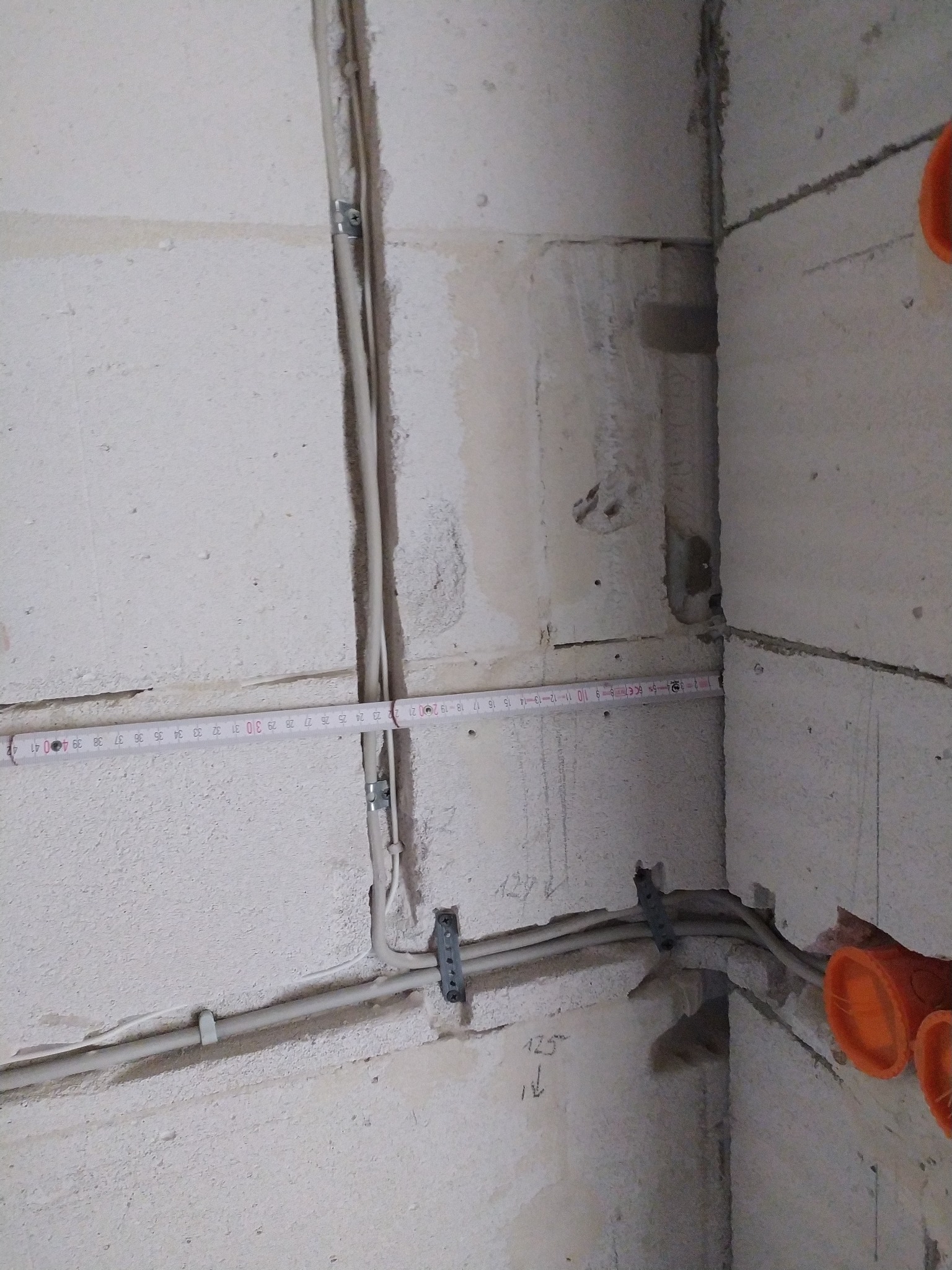 Surowa ściana z zamocowanymi przewodami elektrycznymi, zabezpieczonymi metalowymi uchwytami. Widoczne pomarańczowe rury instalacyjne i miara zwisająca wzdłuż kabla.