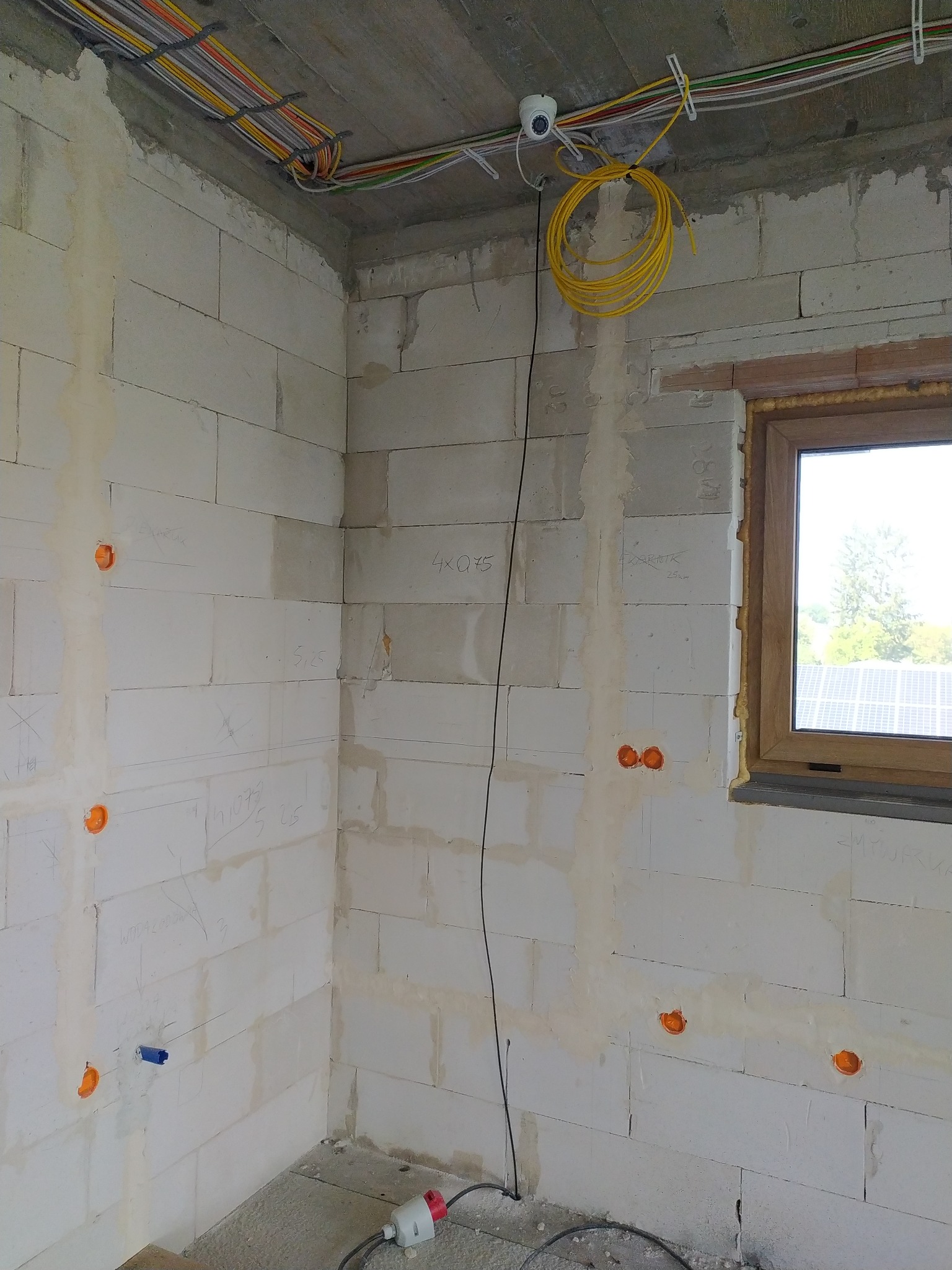 Wnętrze w stanie surowym z widocznymi przewodami elektrycznymi mocowanymi do sufitu, puszkami instalacyjnymi w ścianach i instalacją kamery. Widoczne okno z widokiem na panele słoneczne.
