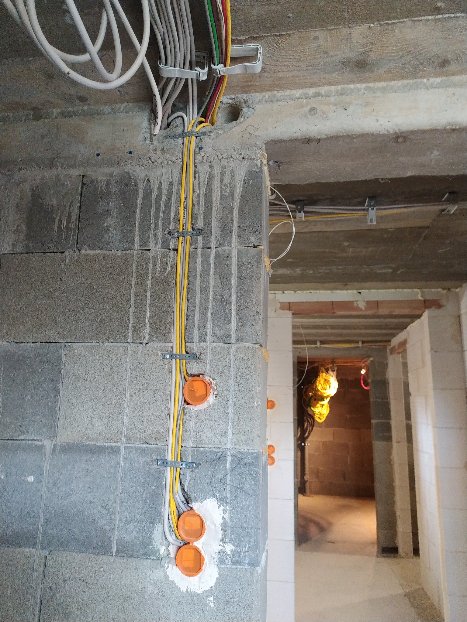 Instalacja elektryczna w surowym wnętrzu: kable w peszlach mocowane do ściany z bloczków betonowych, widoczne puszki instalacyjne i przewody pod sufitem.