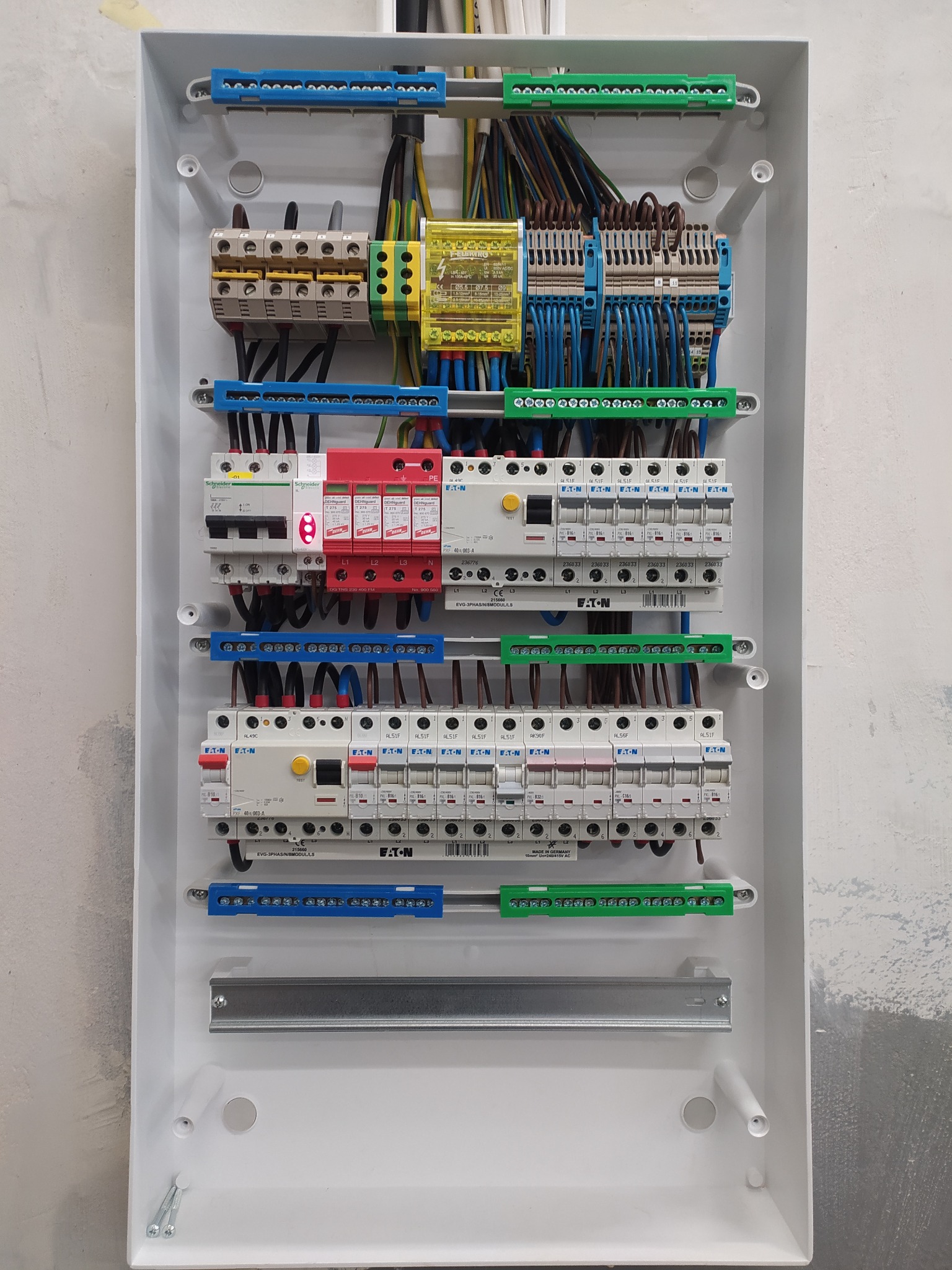 Rozdzielnica elektryczna w białej obudowie, widok z bliska. Widoczne zabezpieczenia, przewody i listwy zaciskowe w kolorach niebieskim, zielonym, żółtym i czerwonym.