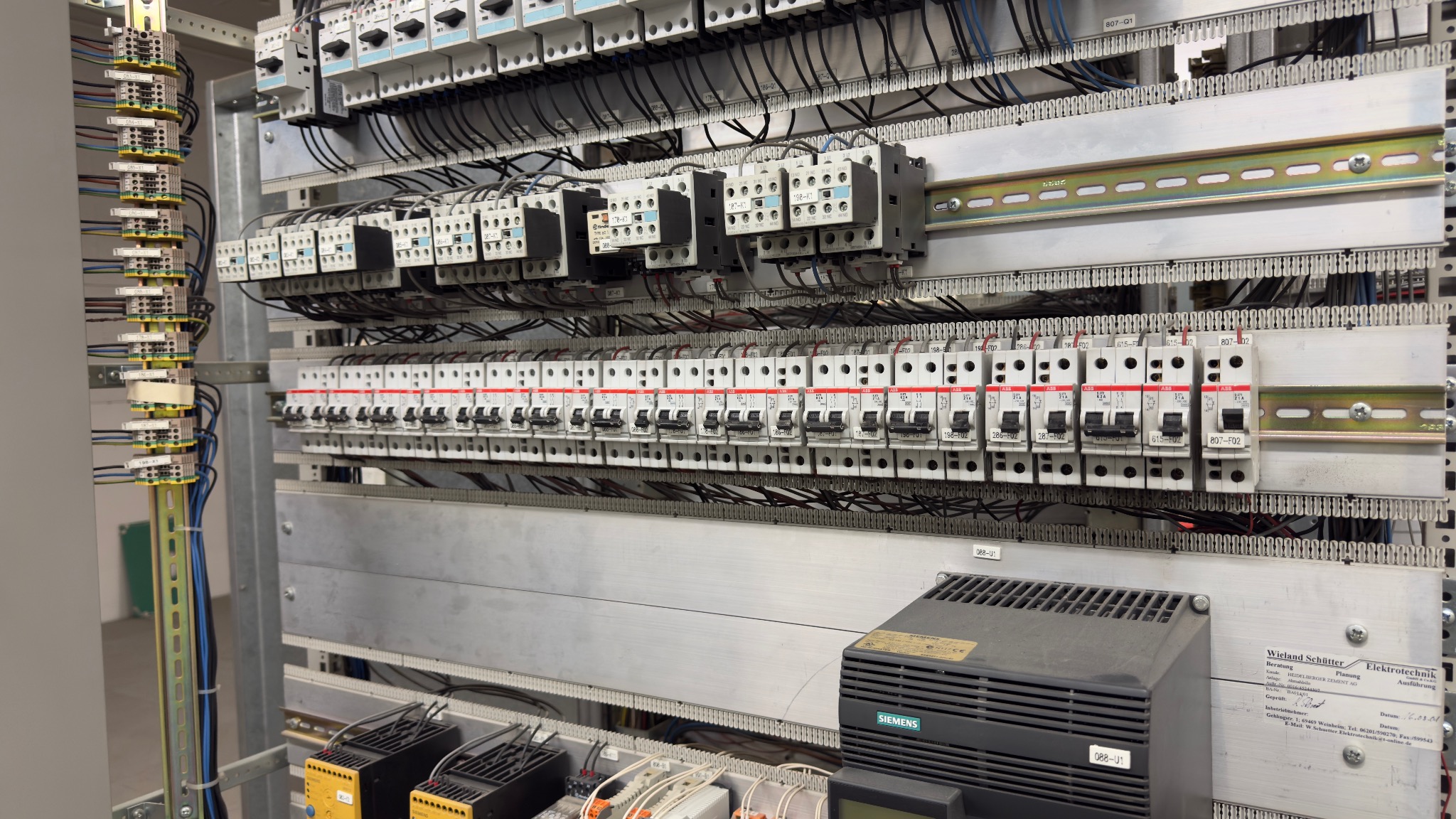 Rozbudowana szafa sterownicza z licznymi bezpiecznikami, stycznikami i okablowaniem. Widoczne elementy marki Siemens i ABB. Precyzyjny montaż w szarej obudowie.