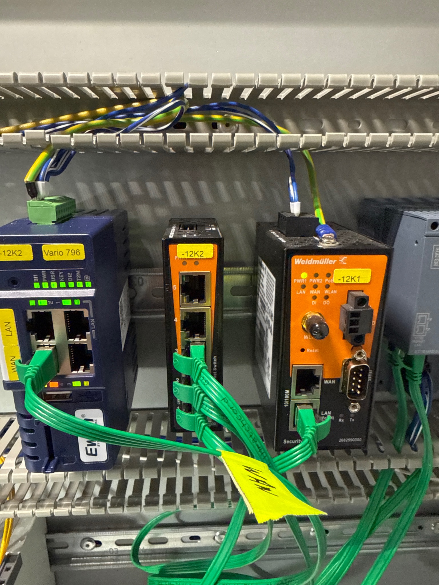 Panel sterowniczy z zamontowanymi modułami sieciowymi Weidmüller i Ewon, połączonymi zielonymi kablami Ethernet. Widoczne oznaczenia i diody LED.