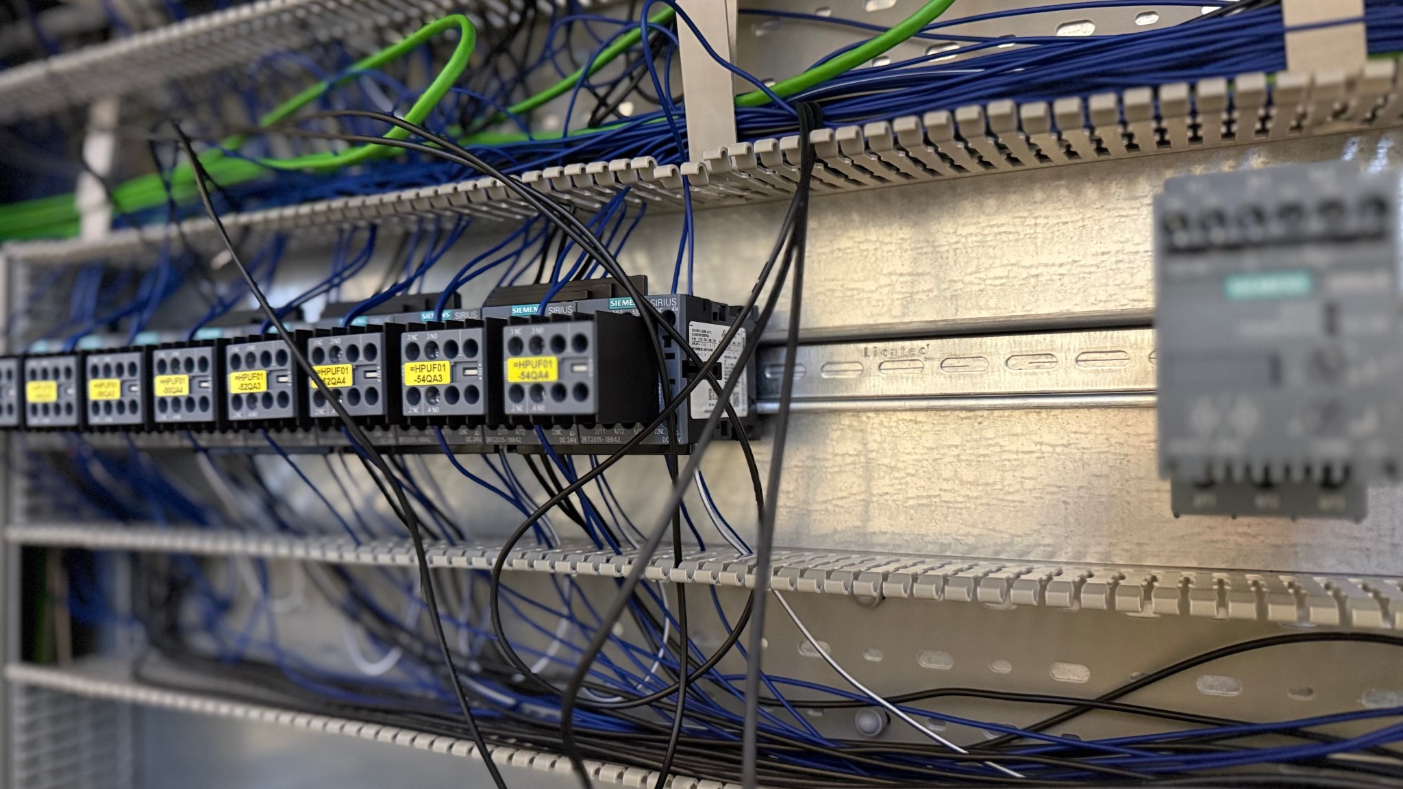 Szczegółowe ujęcie szafy sterowniczej z zamontowanymi stycznikami Siemens Sirius, okablowaniem w kolorach niebieskim i zielonym oraz szyną montażową.