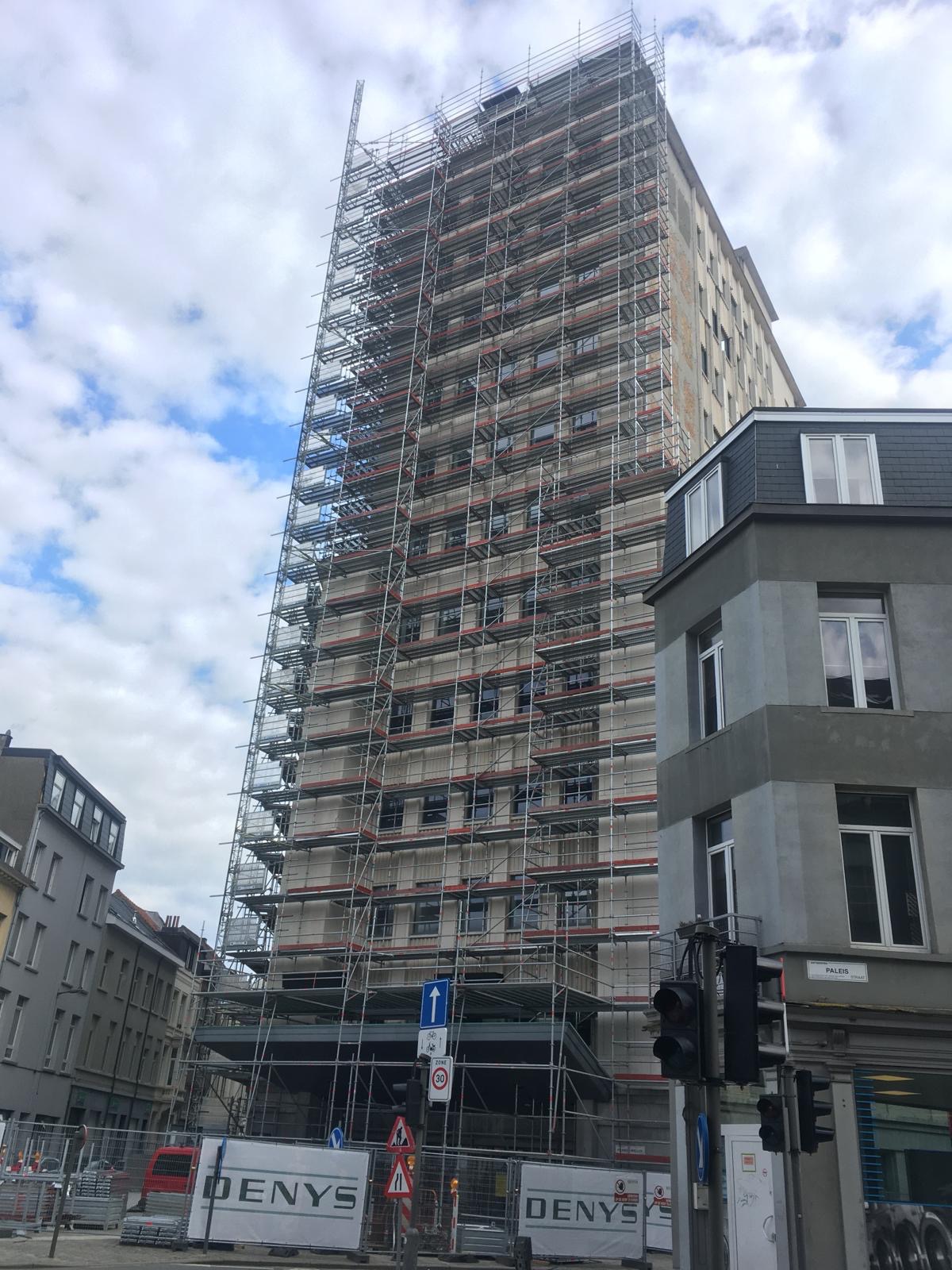 Wysoki budynek w trakcie remontu, całkowicie otoczony rusztowaniami. Widoczne znaki drogowe i fragmenty innych budynków w tle. Ujęcie z dołu, pochmurne niebo.