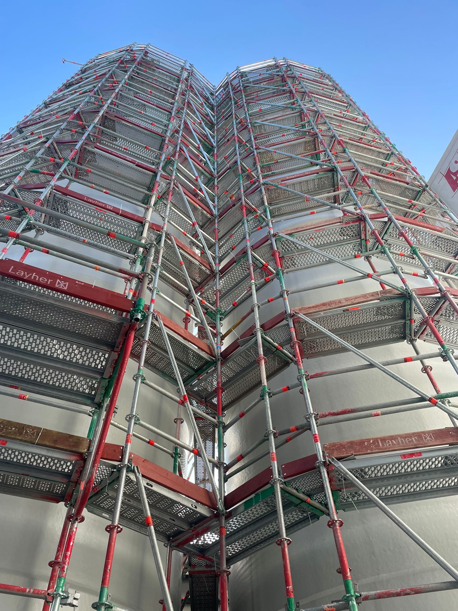 Skomplikowana konstrukcja rusztowania Layher, wznosząca się ku błękitnemu niebu, widziana z dołu. Elementy czerwone, zielone i srebrne tworzą geometryczny wzór.