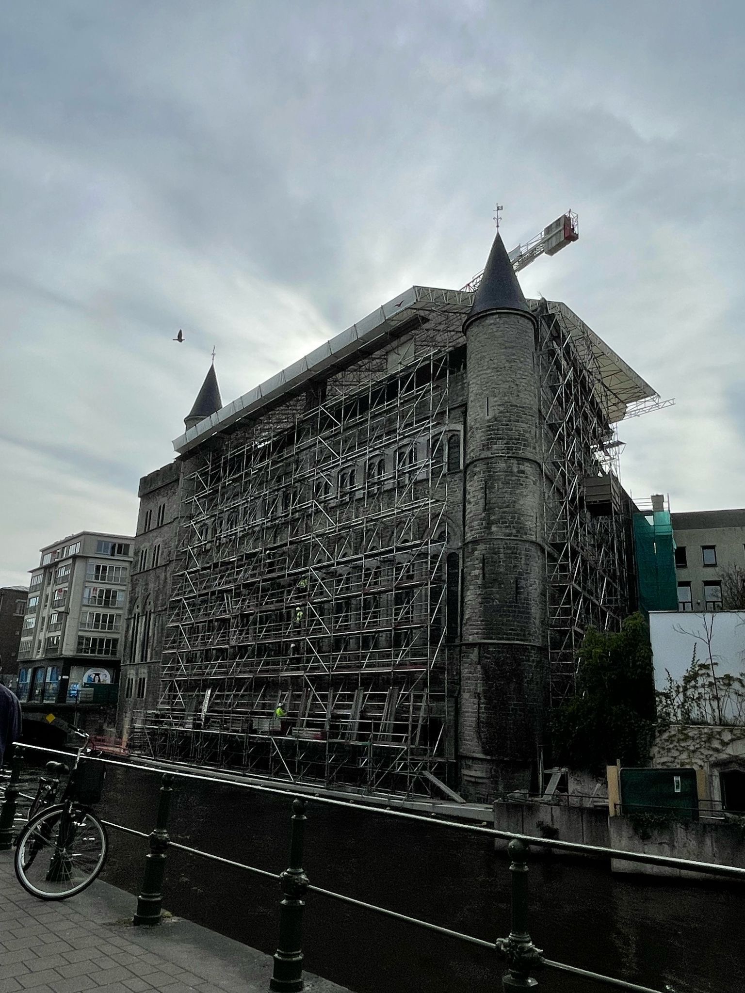 Kamienny budynek w rusztowaniach, widok z dołu, z elementami rzeki i roweru w kadrze. Na szczycie budynku widoczny żuraw. W tle pochmurne niebo.