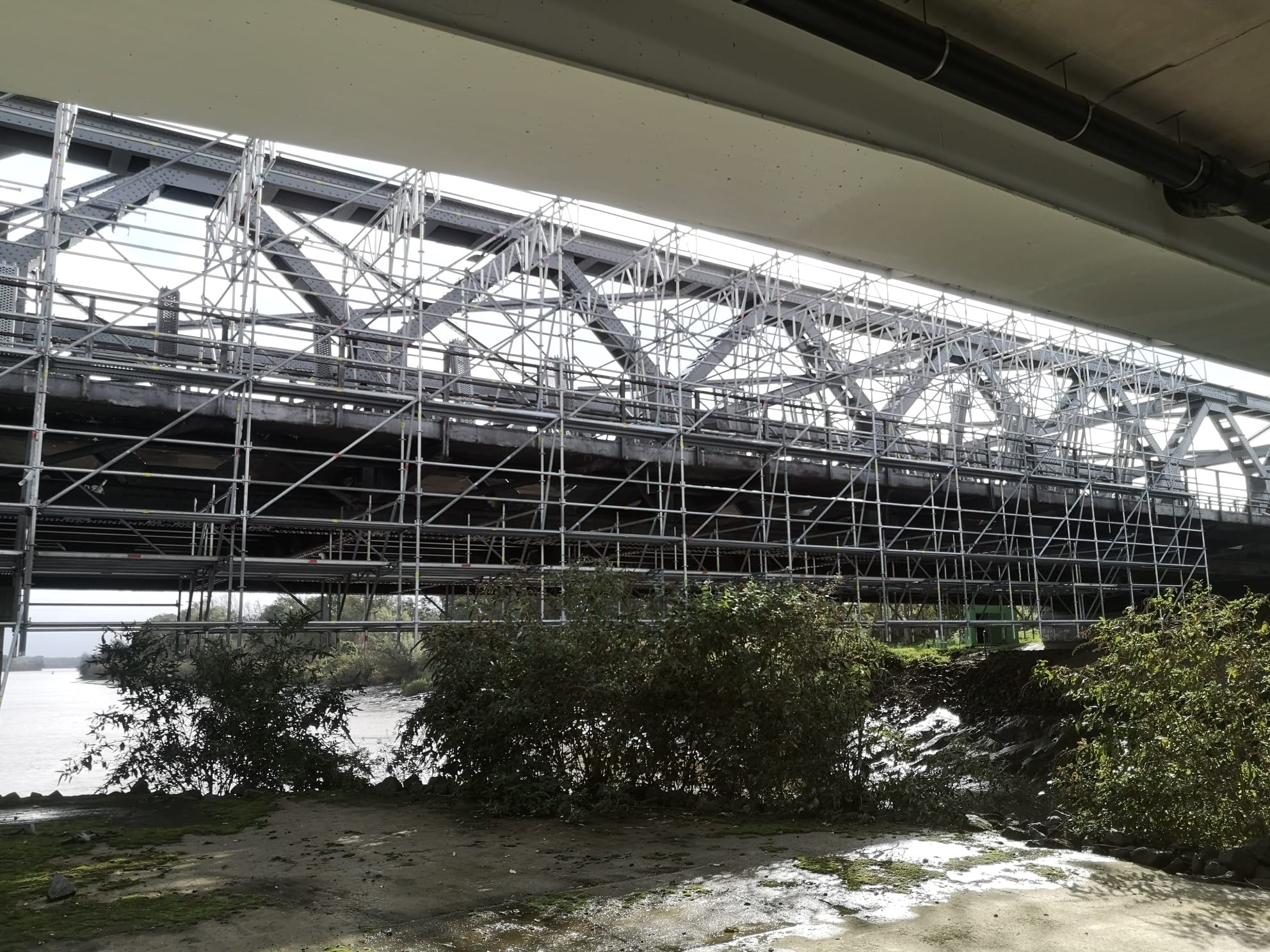 Metalowa konstrukcja mostu w trakcie prac wysokościowych, otoczona rusztowaniami. Widok z dołu, spod mostu, z zielenią w tle.