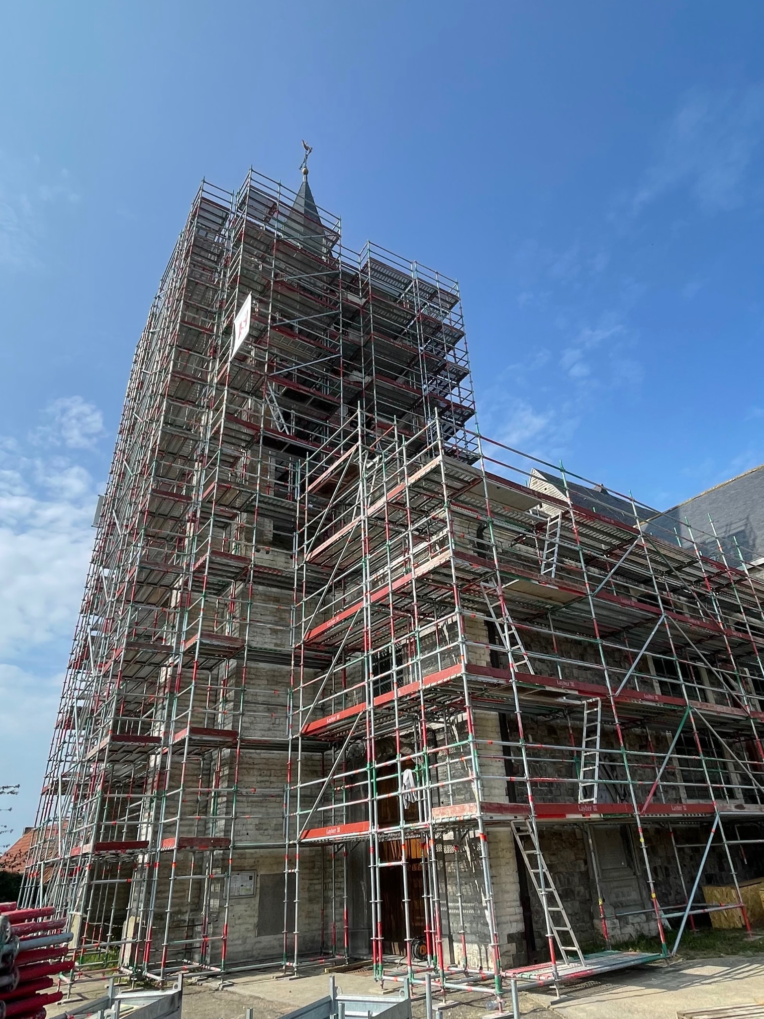 Kościół w rusztowaniach pod błękitnym niebem. Widoczne drabiny i elementy konstrukcyjne rusztowań. Prace renowacyjne na wysokościach.