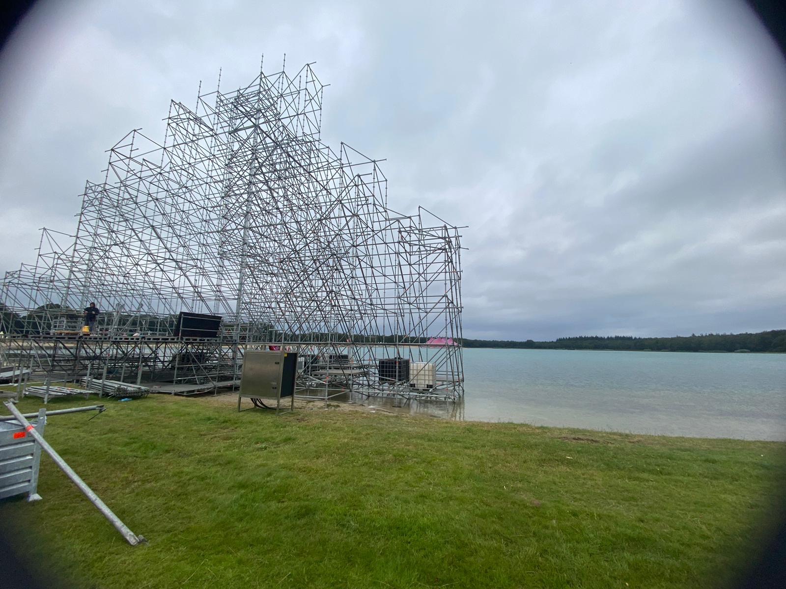 Skomplikowana konstrukcja rusztowania nad jeziorem, widoczna z poziomu trawy, pod pochmurnym niebem. Elementy rusztowania częściowo zanurzone w wodzie.