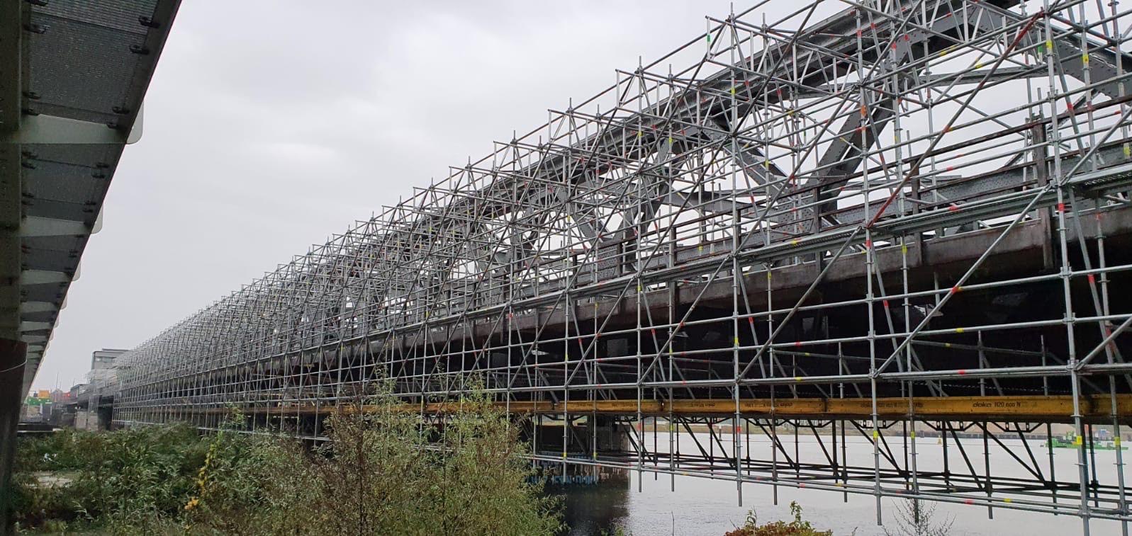 Skomplikowana konstrukcja rusztowań otaczająca stalowy most, widoczna z dołu, z zielenią i wodą w tle, przy pochmurnym niebie. Detaliczna siatka rusztowań.