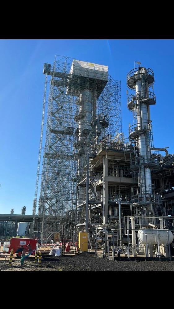 Rozległa instalacja przemysłowa z metalowymi rusztowaniami otaczającymi wysokie kolumny, na tle błękitnego nieba. Widoczne rury, zbiorniki i elementy konstrukcyjne.