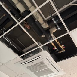 Instalacja rur i zaworów pod sufitem podwieszanym, widoczny klimatyzator kasetonowy. Różne materiały rur: czarne izolowane, białe PCV, miedziane z zaworami.