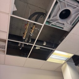 Odsłonięty sufit podwieszany z widocznymi rurami instalacji hydraulicznej, wentylacją i oświetleniem. Widoczny zegar na ścianie.